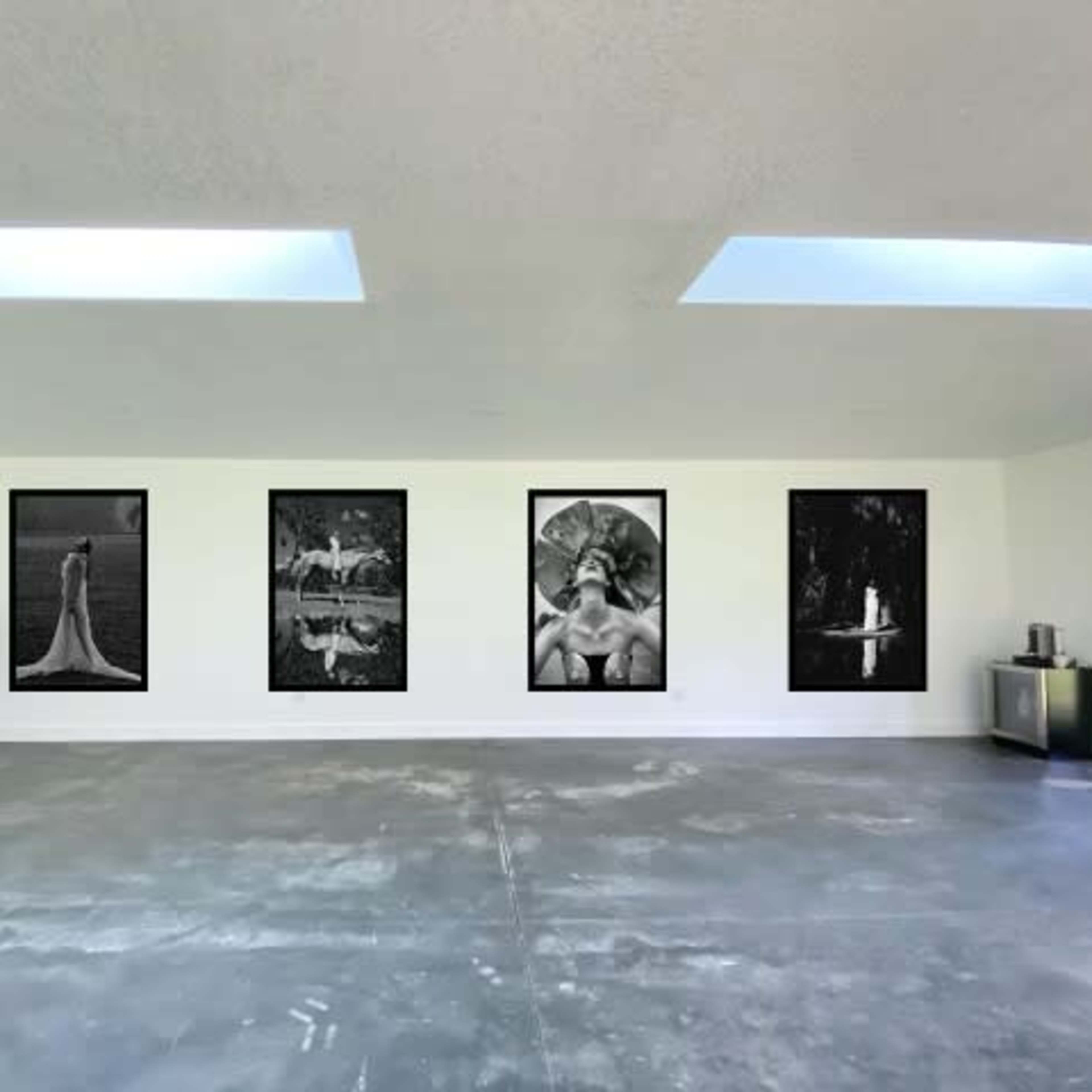 A spacious, minimalist art gallery featuring four black-and-white photographs on the walls, with clothing displayed on a rack in the corner.