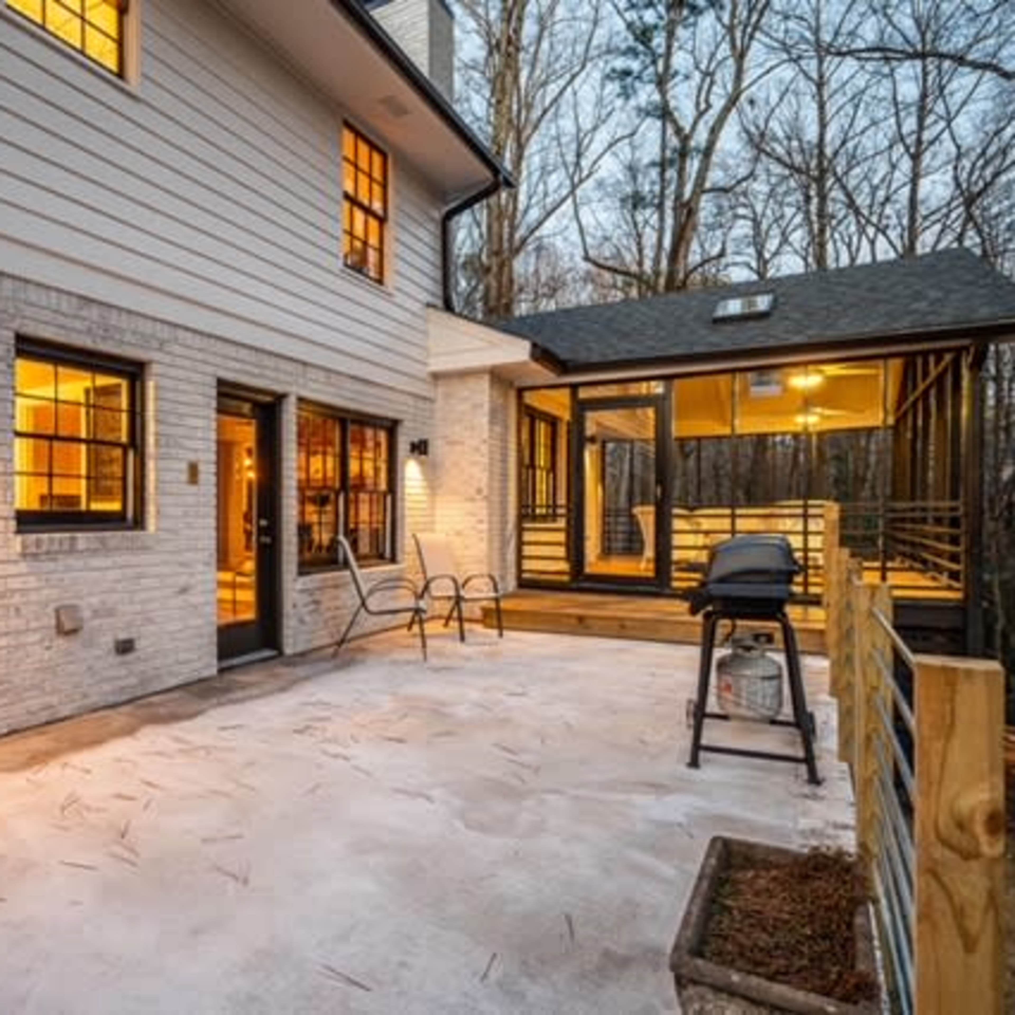 A patio area adjacent to a two-story house, featuring a grill and surrounded by trees.