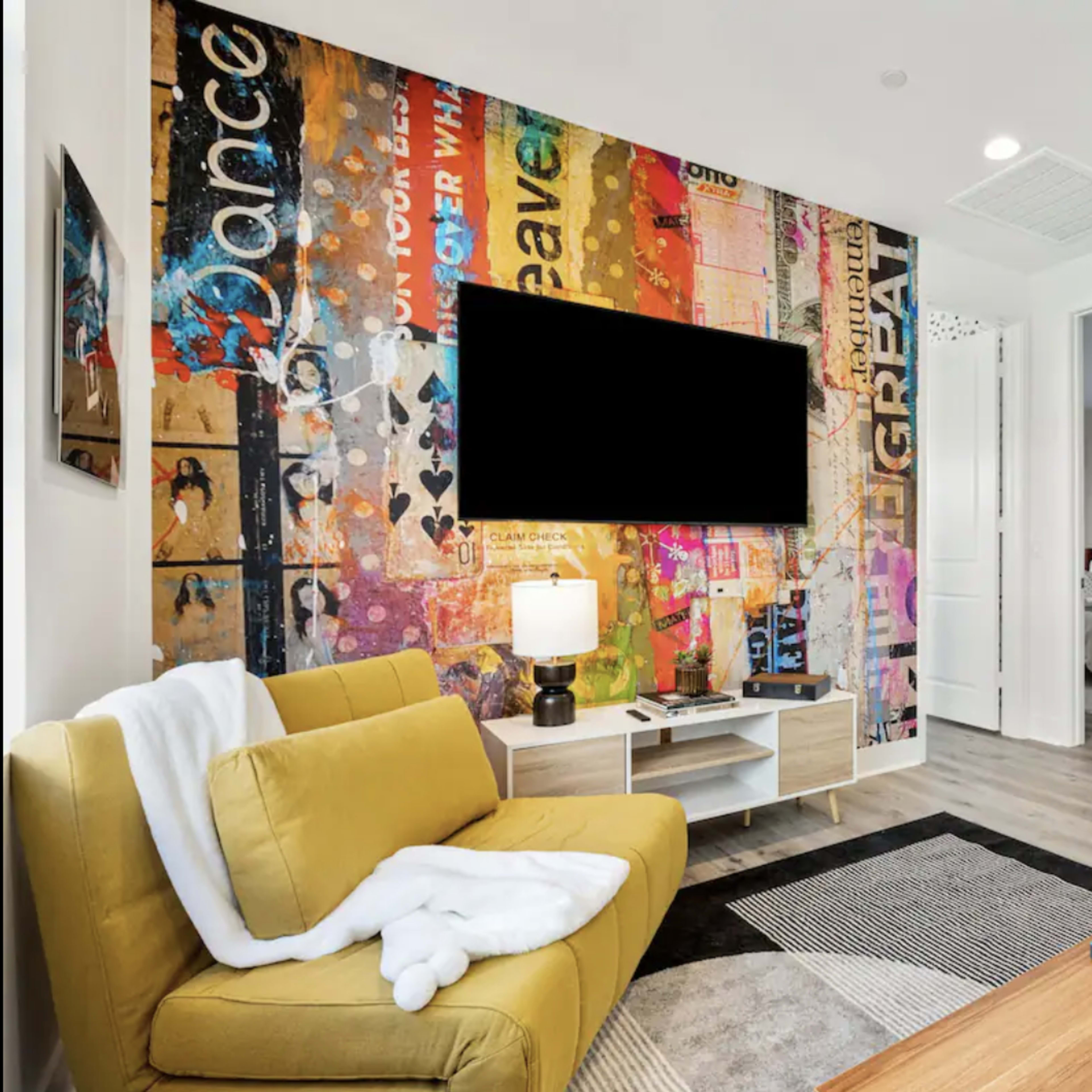 A living room features a yellow sofa, a coffee table with a vinyl record, and a wall mural with colorful graphics, alongside a mounted television.
