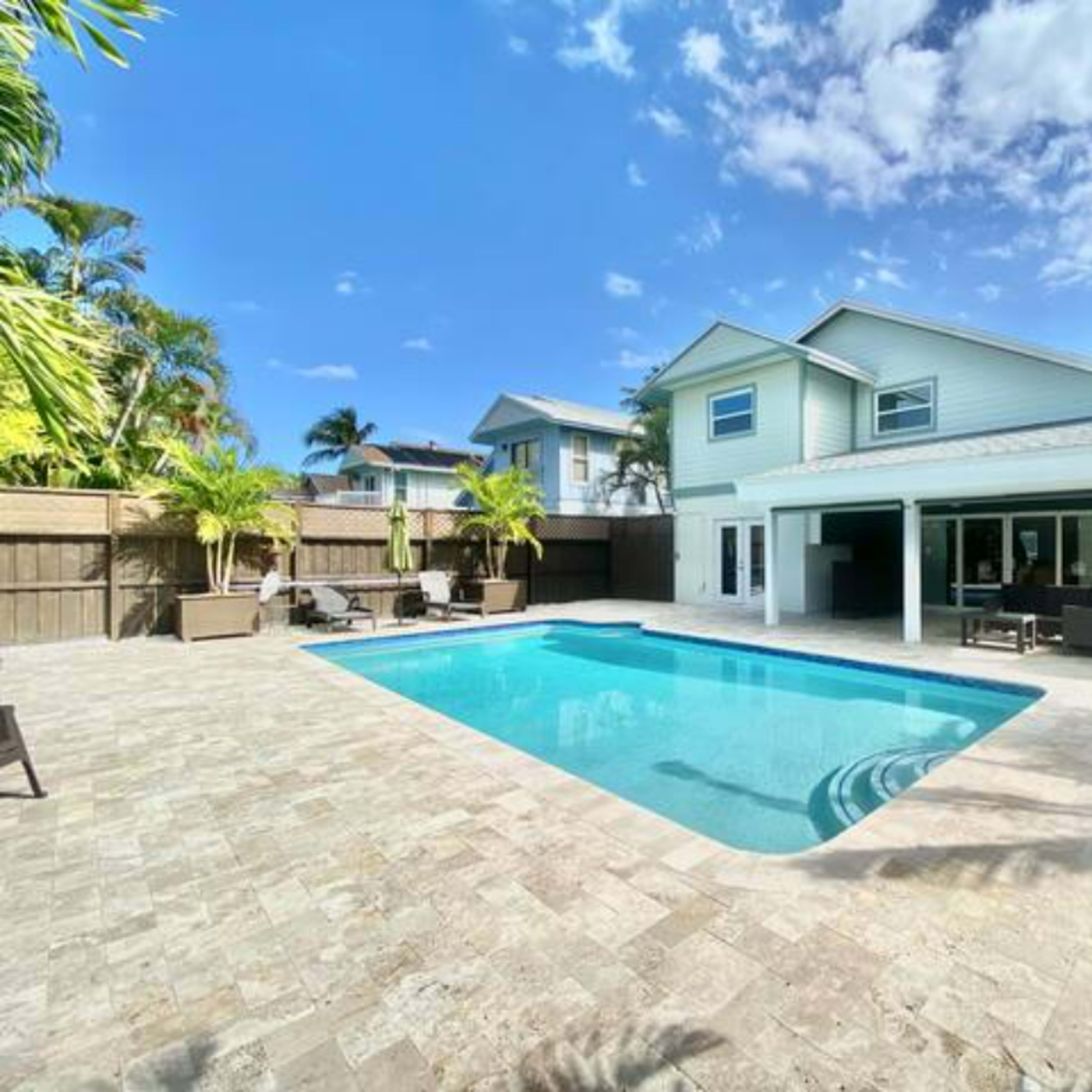 A backyard with a swimming pool, surrounded by stone pavement and tropical landscaping, with a two-story house in the background.