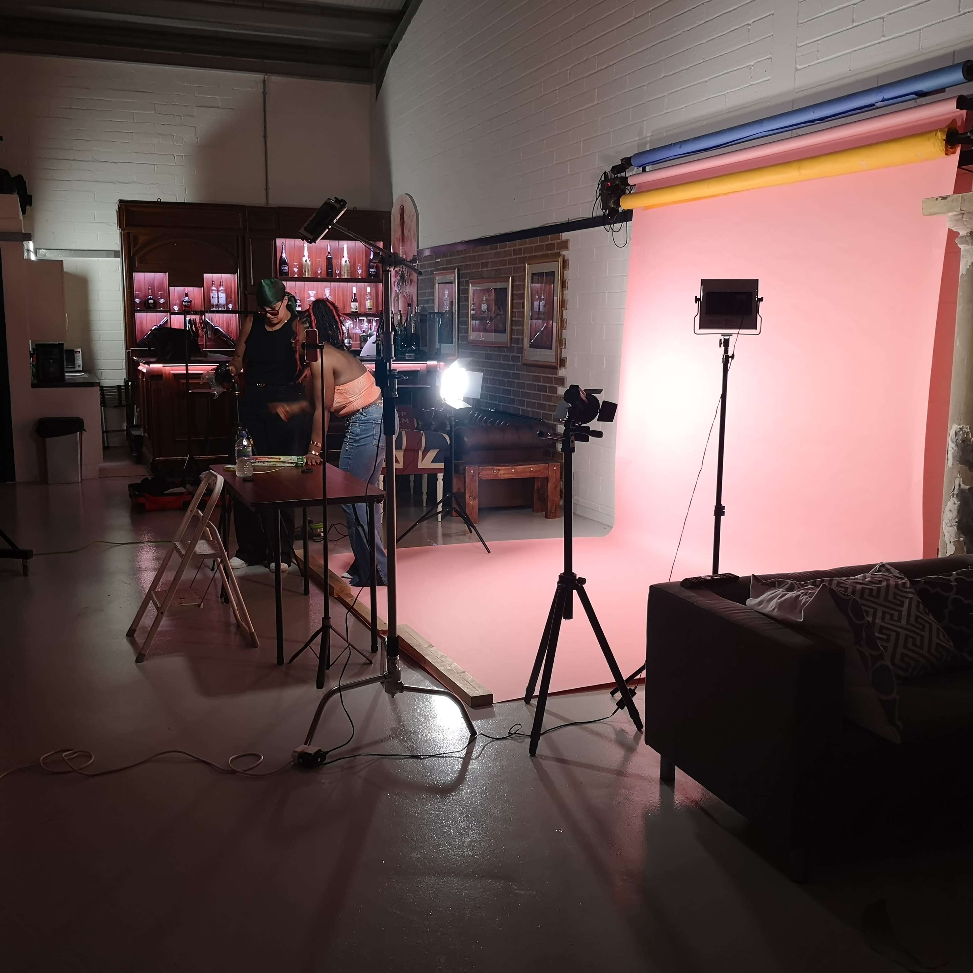 A photographer sets up a studio with a pink backdrop, lighting equipment, and a table in a room featuring a bar and furniture.