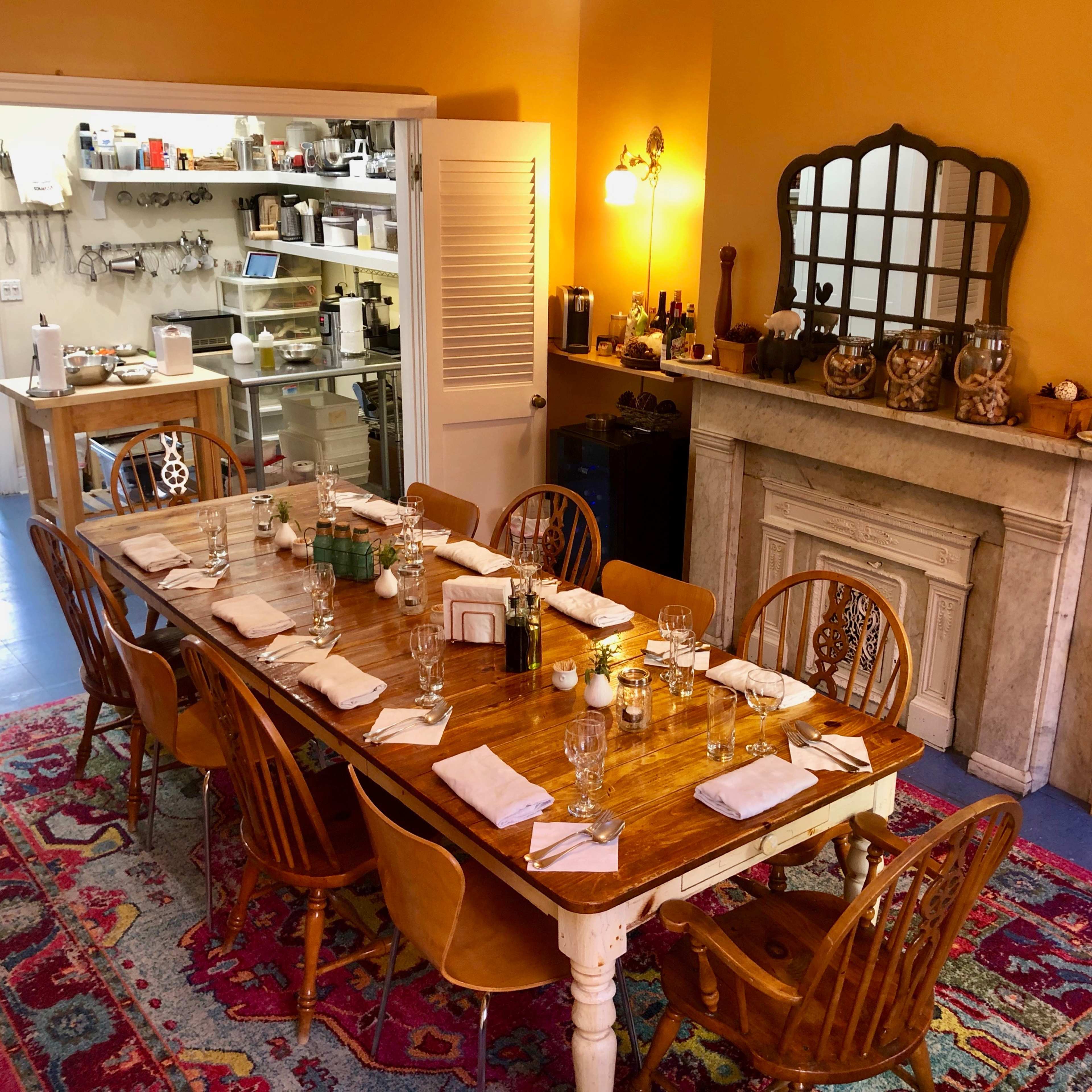 A long wooden dining table is set with plates and glasses in a warmly lit dining room, with a kitchen area visible in the background.