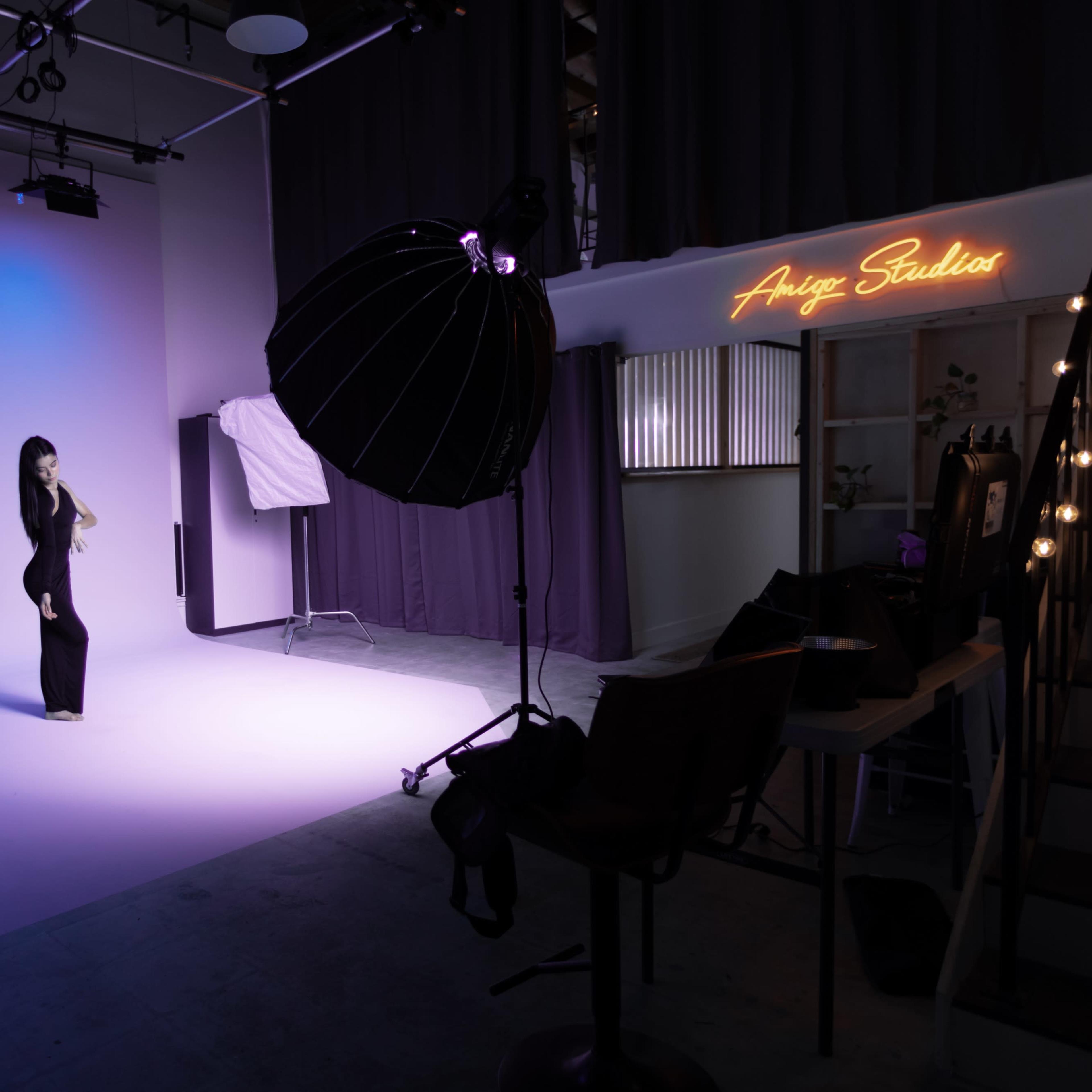 A model poses against a gradient blue backdrop in a photography studio, with studio lights and equipment arranged nearby.