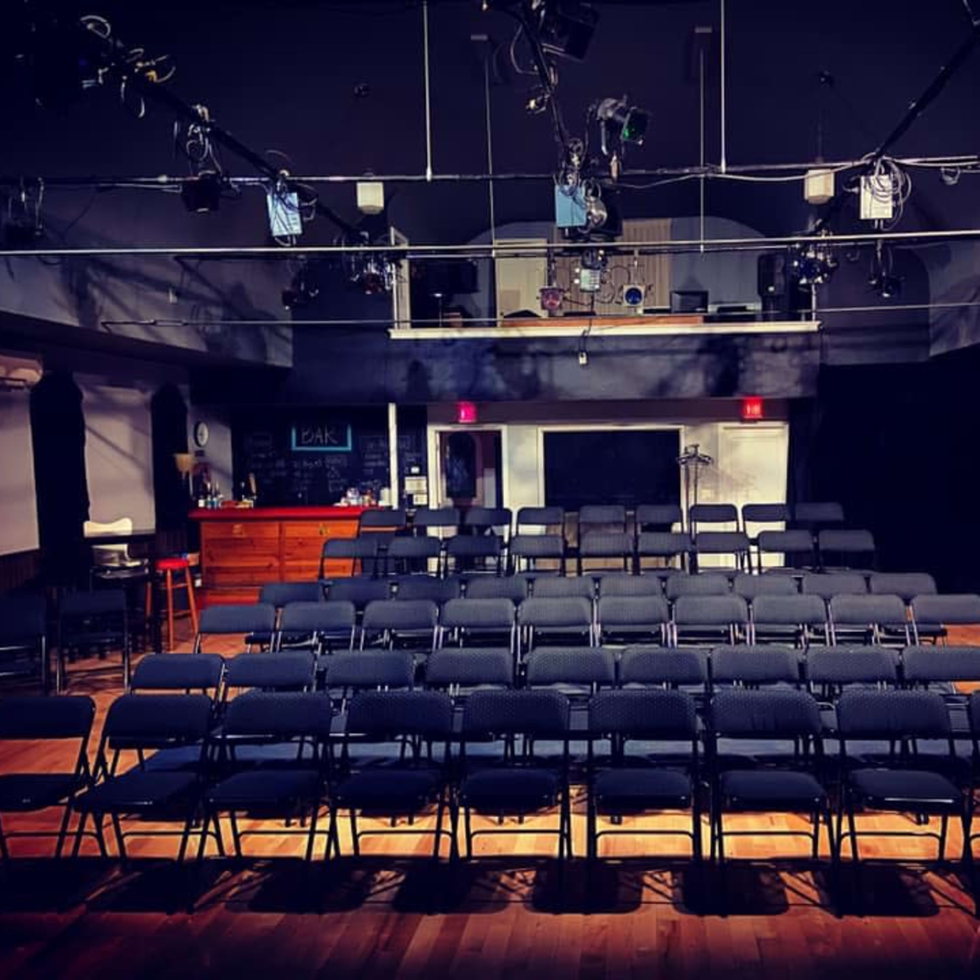A theater setup with rows of black folding chairs facing a stage, illuminated by overhead stage lights.