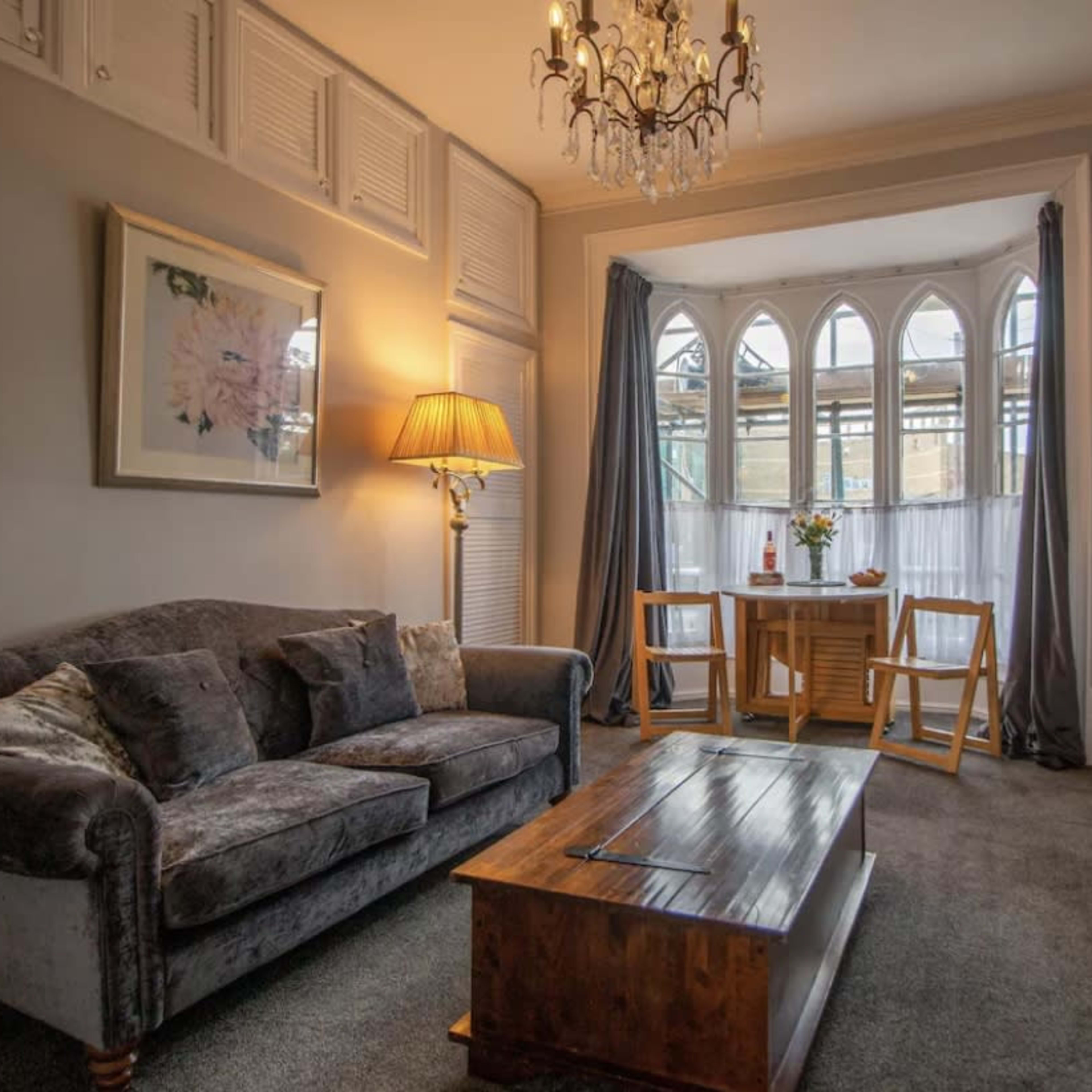 A cozy living room with a gray sofa, a wooden coffee table, and a dining area featuring two chairs and a small table next to large arched windows.