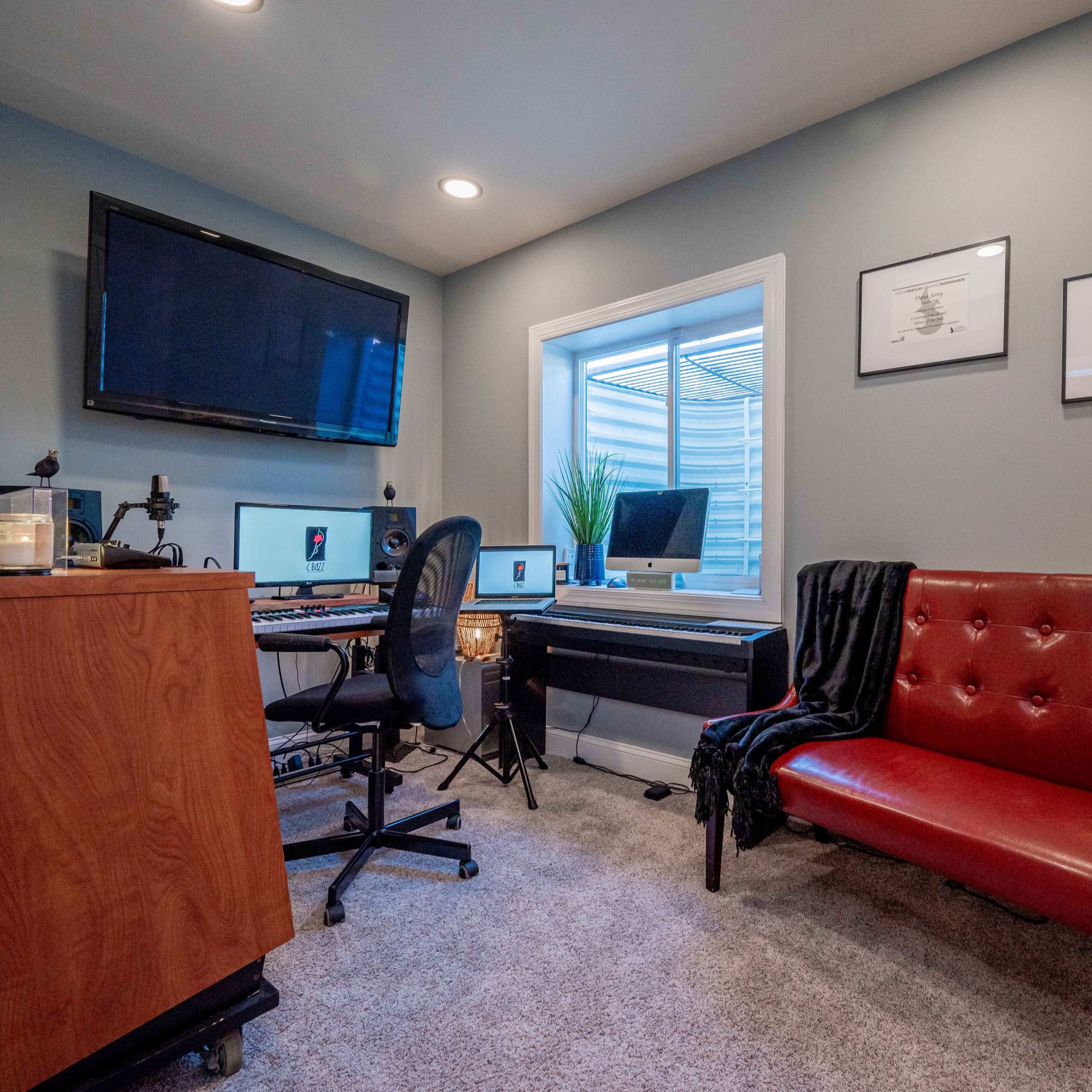 A small home office with a desk, computer, keyboard, a red couch, and a window revealing a plant outside.