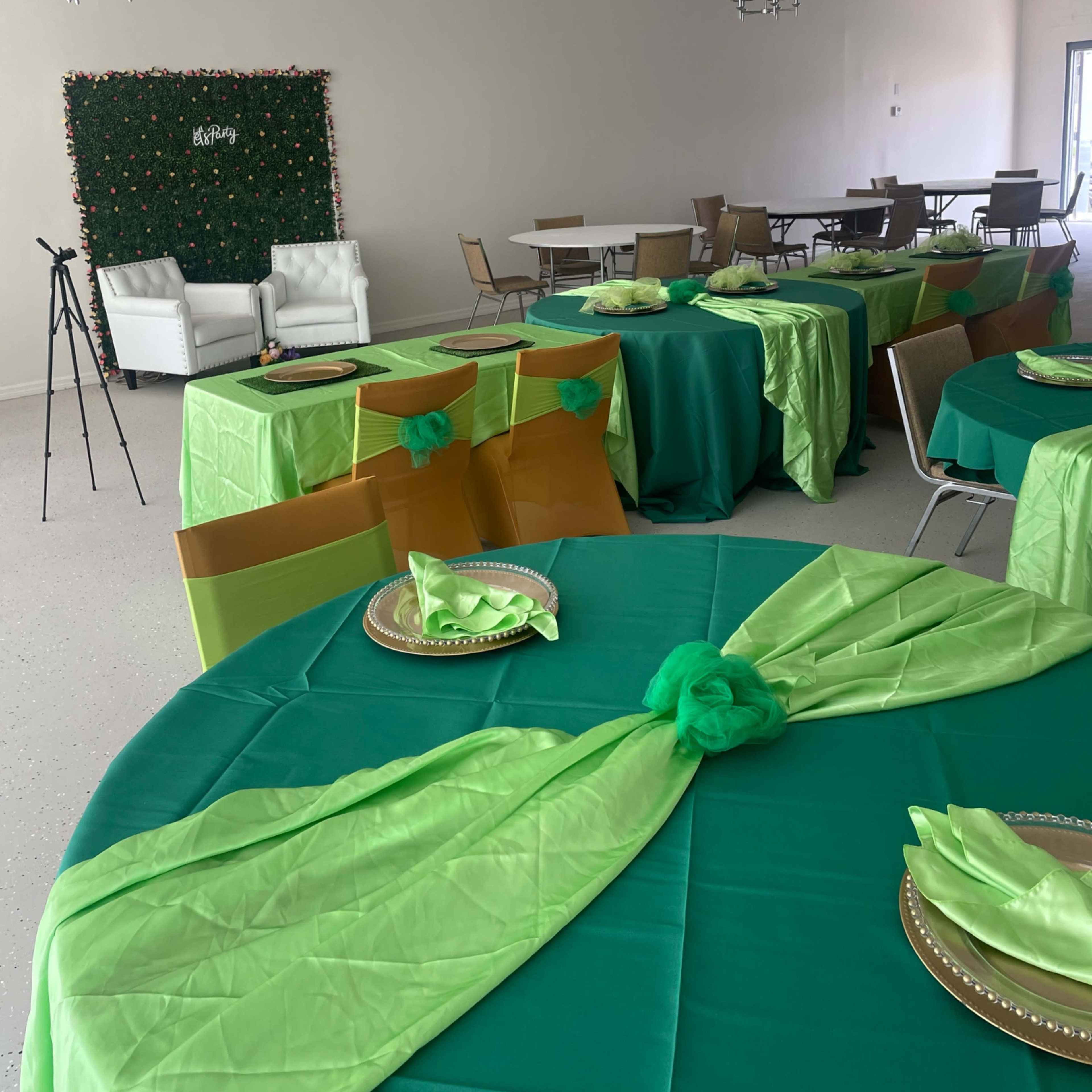 A spacious event room with green and gold tablecloths draped over several tables, neatly set with plates and napkins, and featuring white chairs in the background.