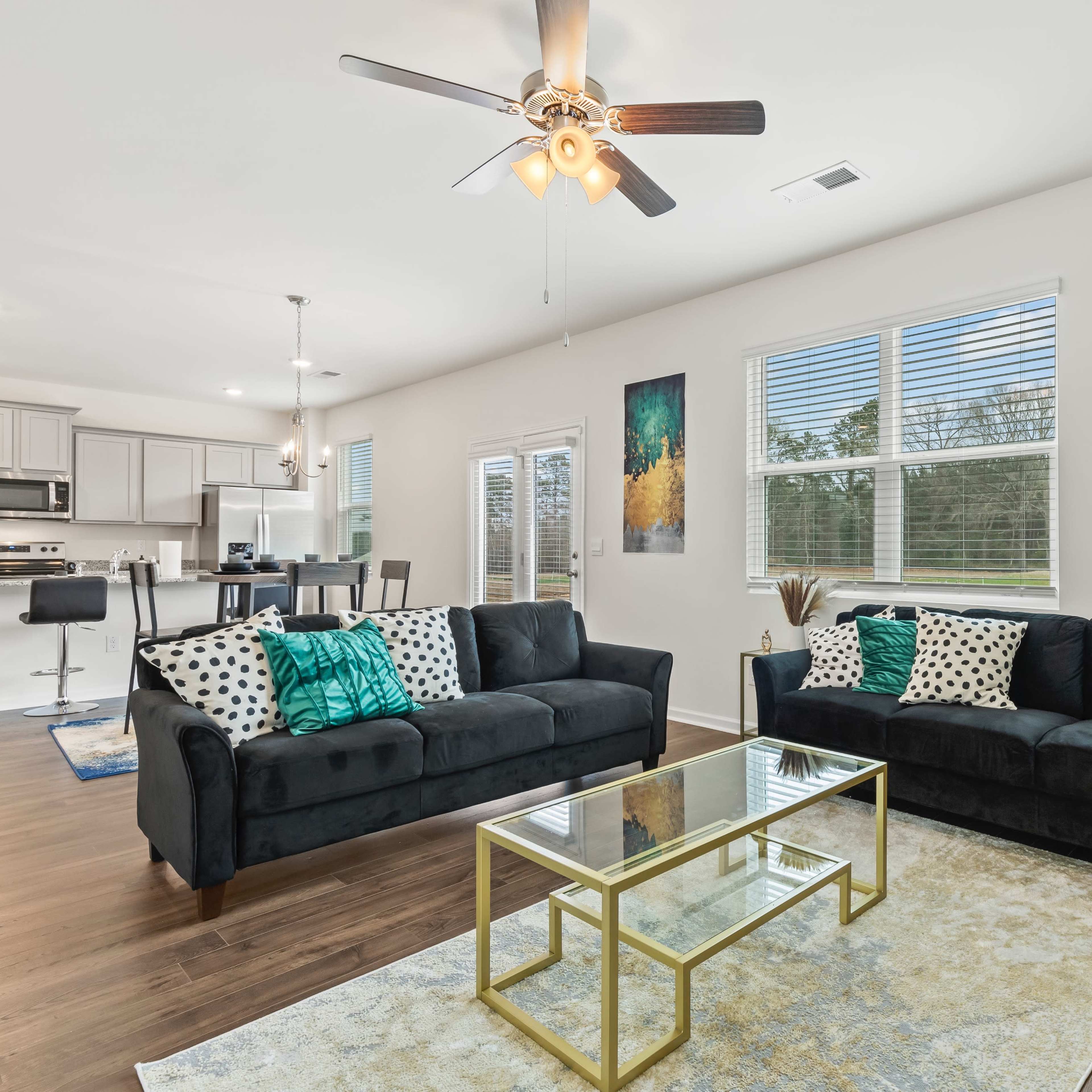 A modern living room with two dark sofas, a glass coffee table, and decorative artwork on the walls, illuminated by natural light from large windows.