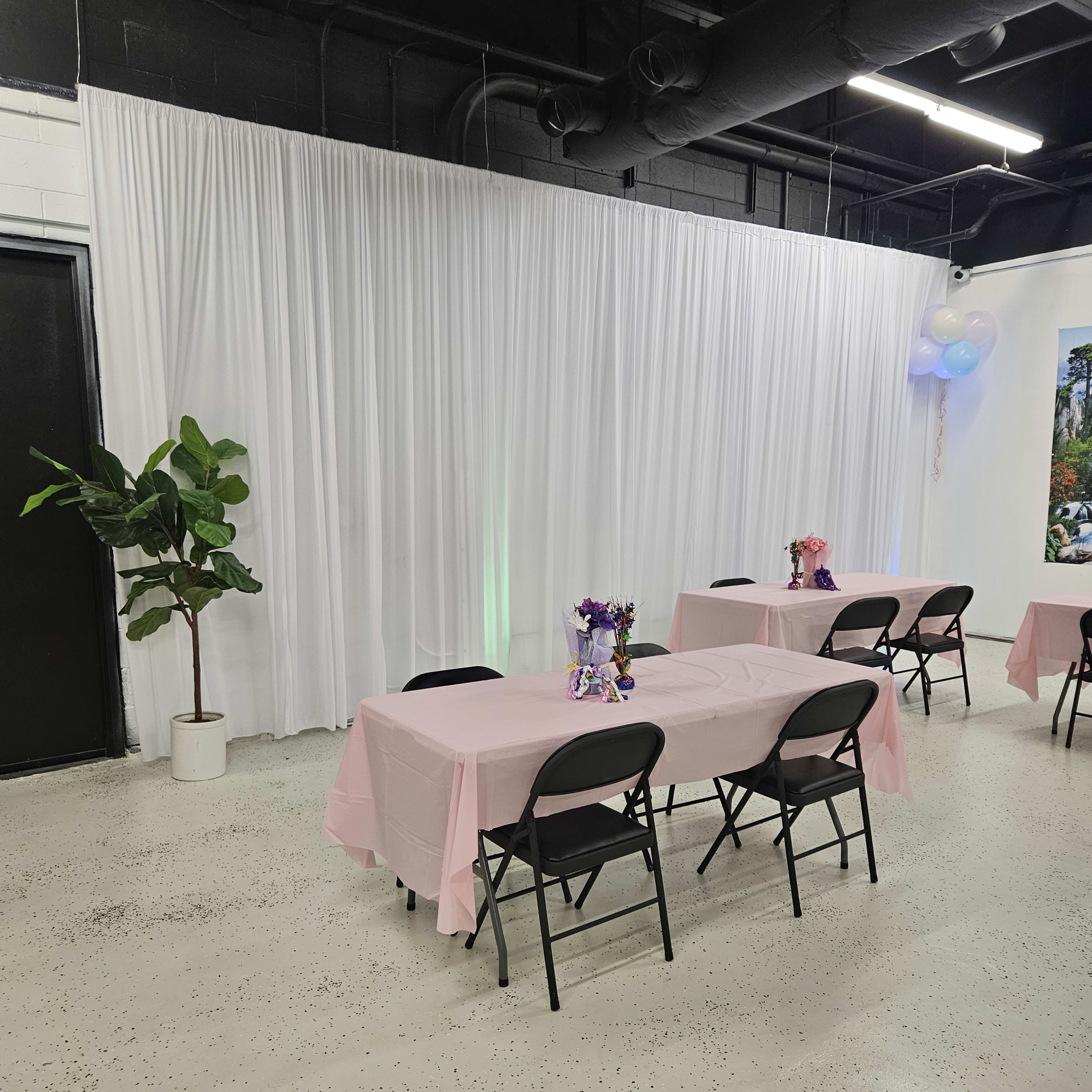 A room with pink-covered tables and a large white backdrop, featuring potted plants and decorative centerpieces.