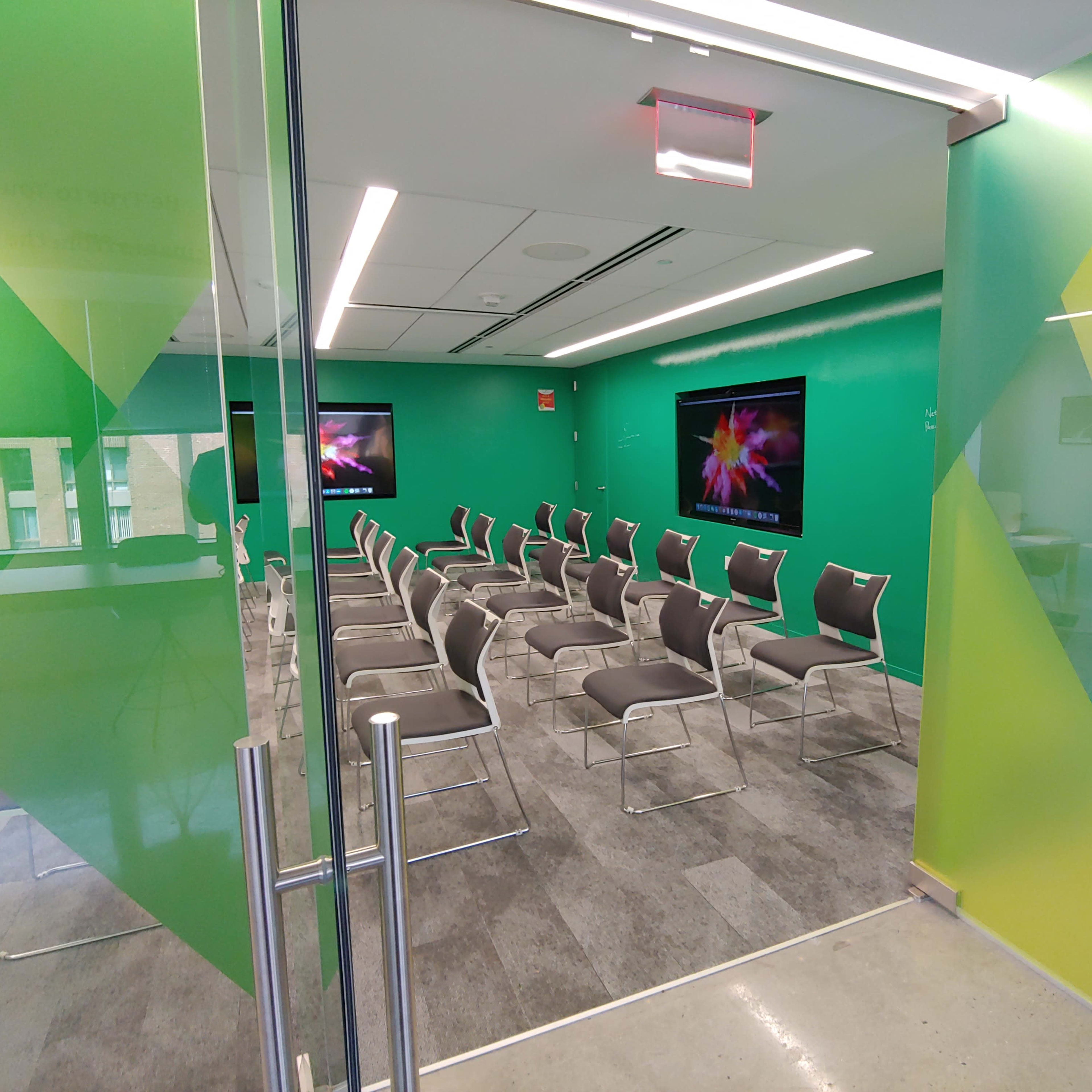 A modern conference room with green walls, two screens, and several rows of gray chairs arranged for a presentation.