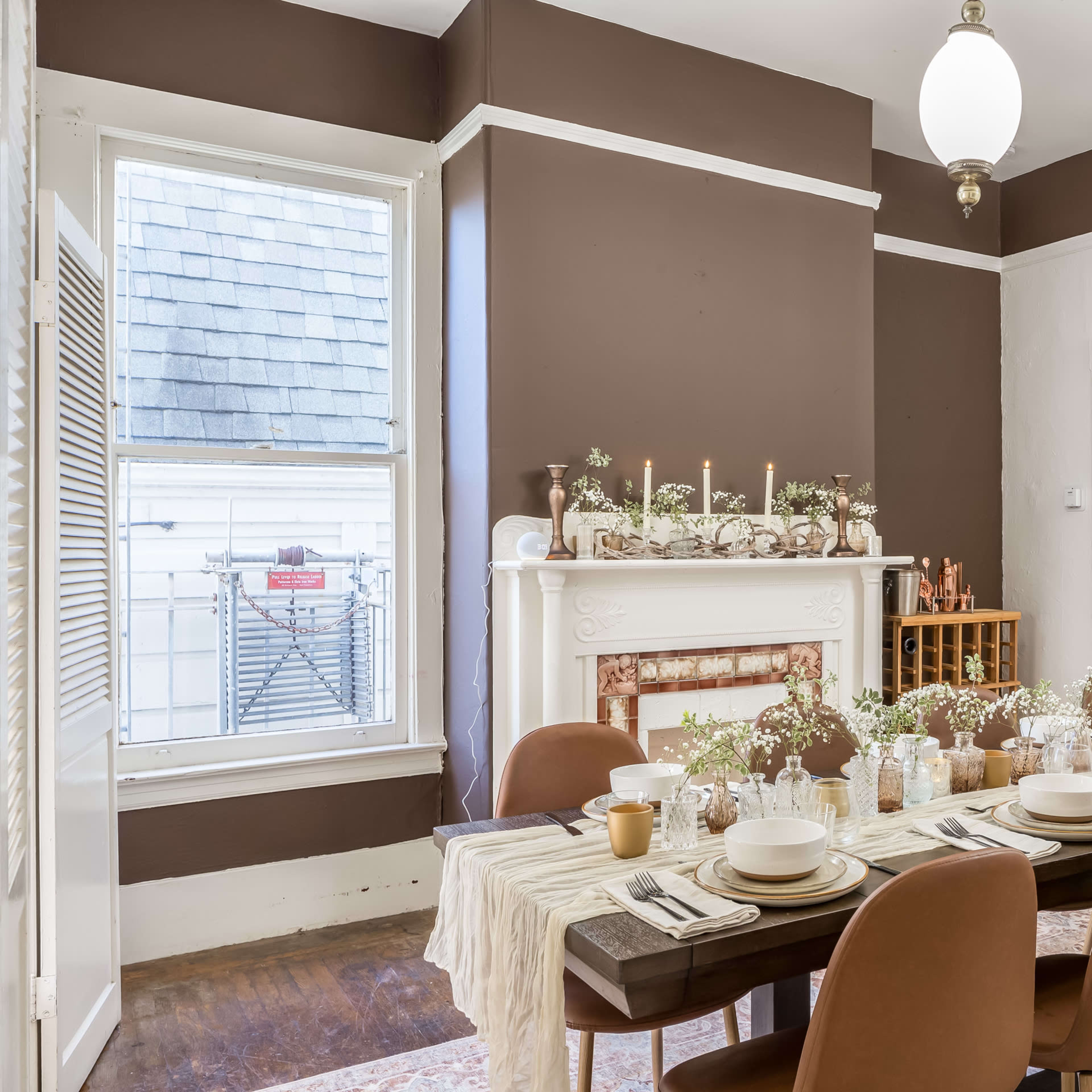 A dining room features a long table set for a meal, with a decorative centerpiece and candles, against a backdrop of brown walls and a white fireplace.