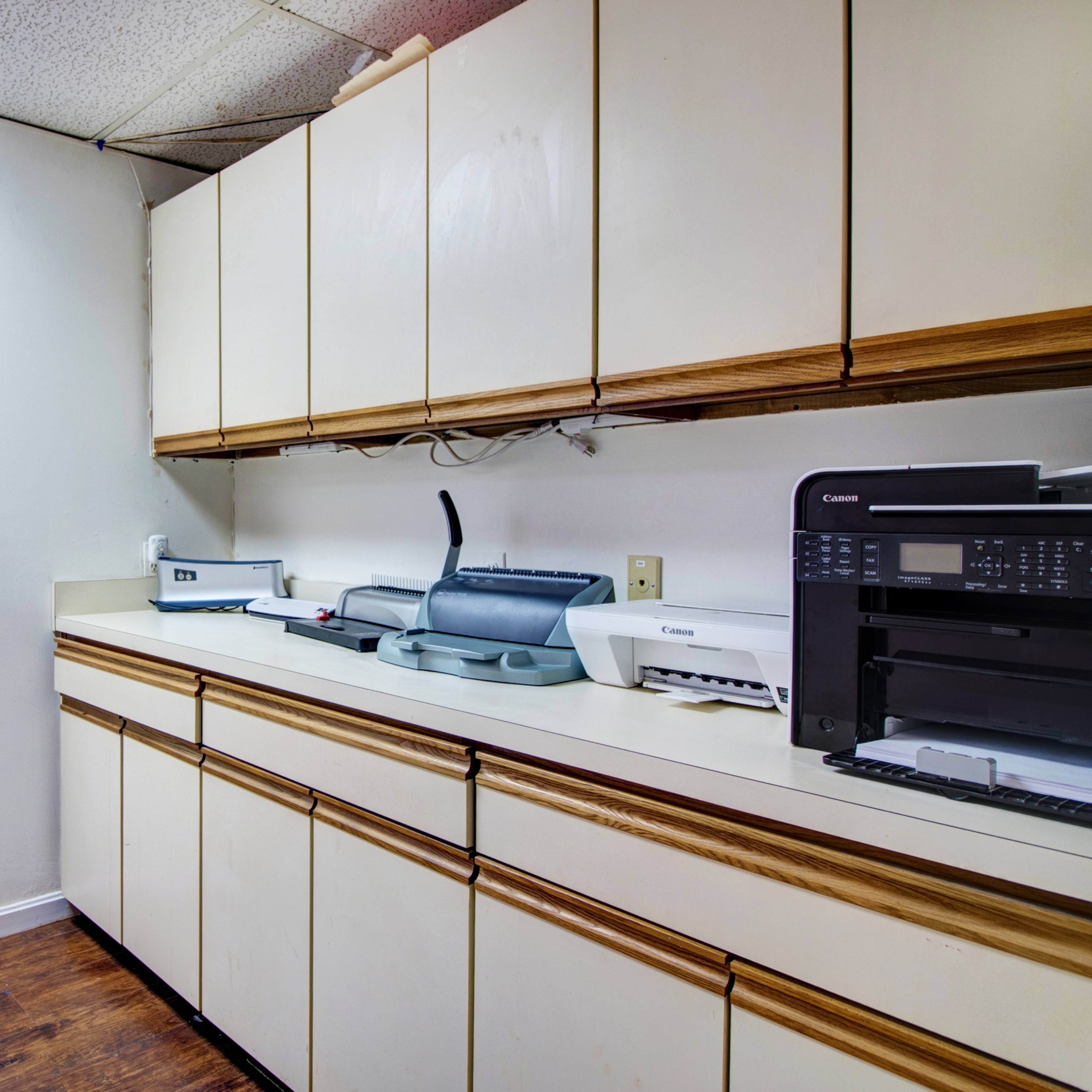 A narrow hallway leads to a kitchen area with beige cabinets, a countertop, and various office equipment, including printers and a scanner.