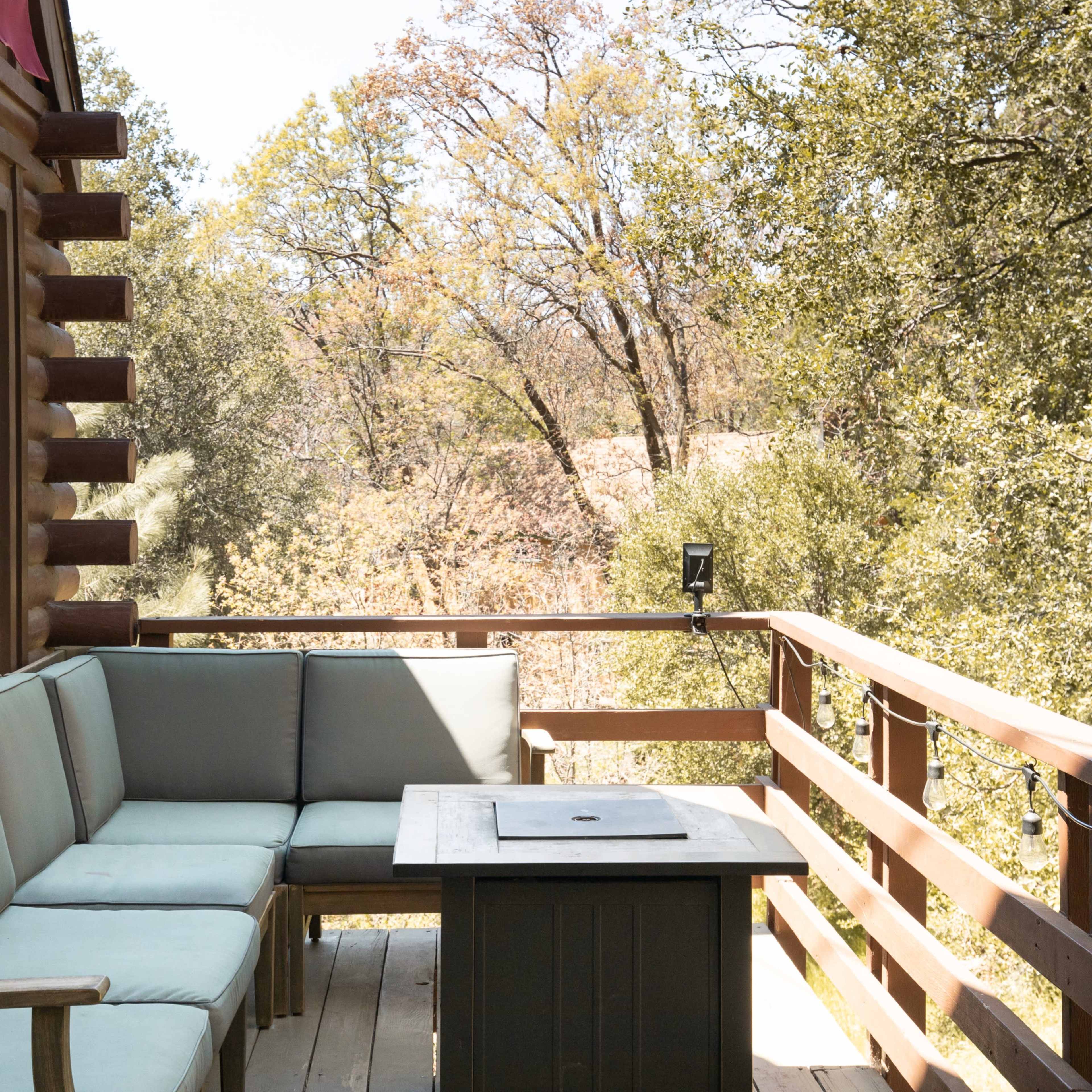 A wooden deck with a green cushioned seating area and a small table overlooks a natural landscape with trees.