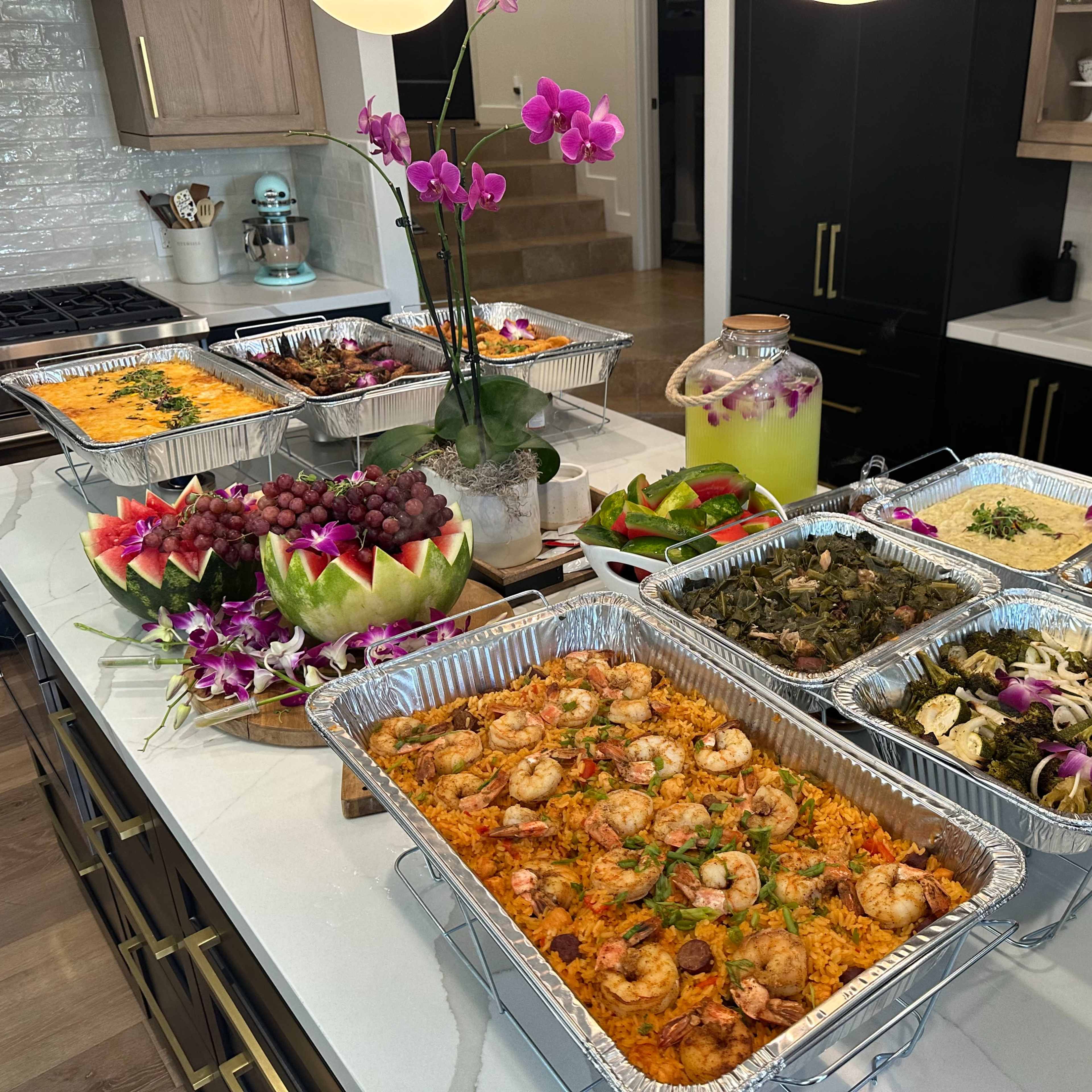 A large buffet of various prepared dishes, including shrimp, rice, and vegetables, is displayed on a kitchen island surrounded by fresh fruit and floral decorations.