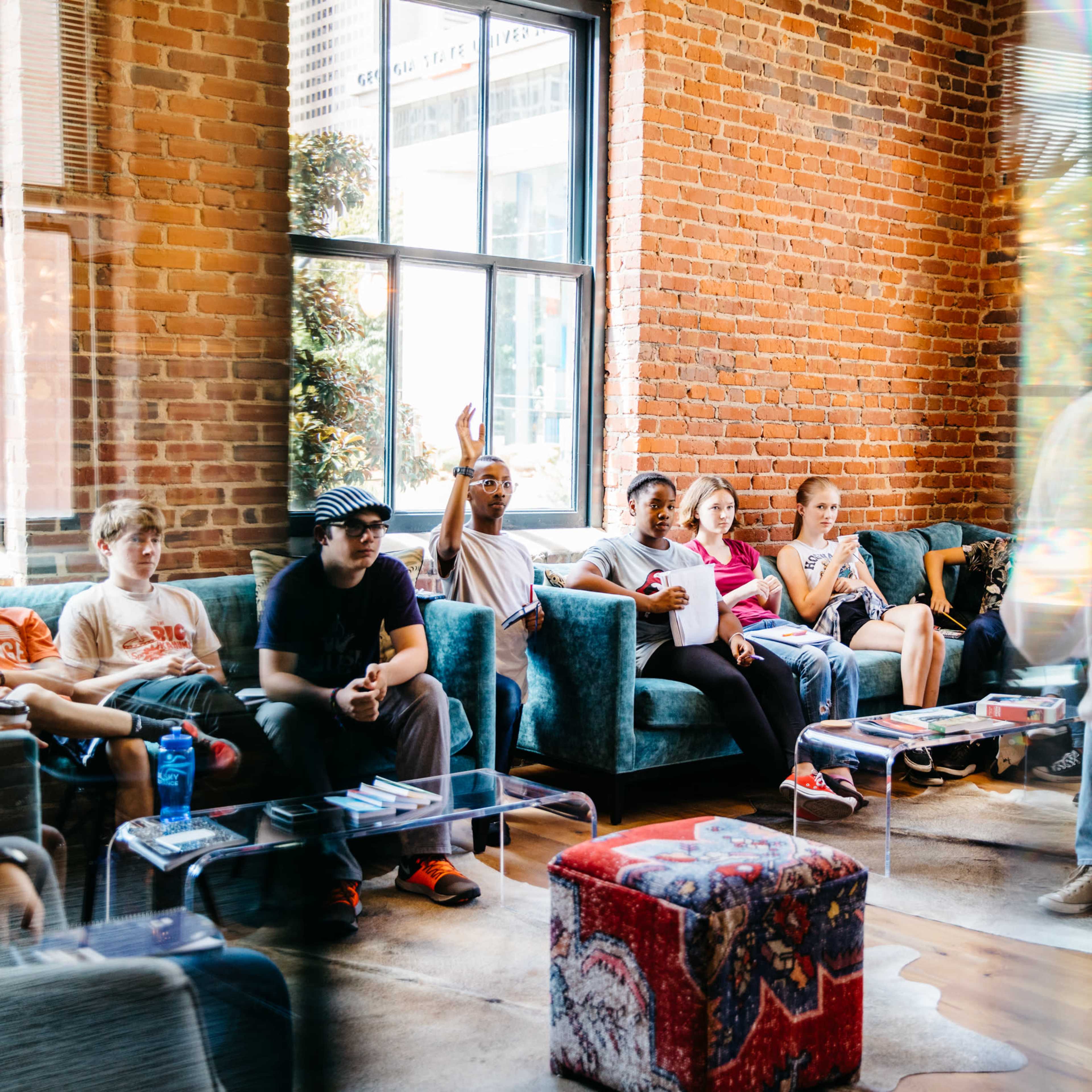 A group of people sits on sofas in a room with brick walls while someone stands and speaks to them.
