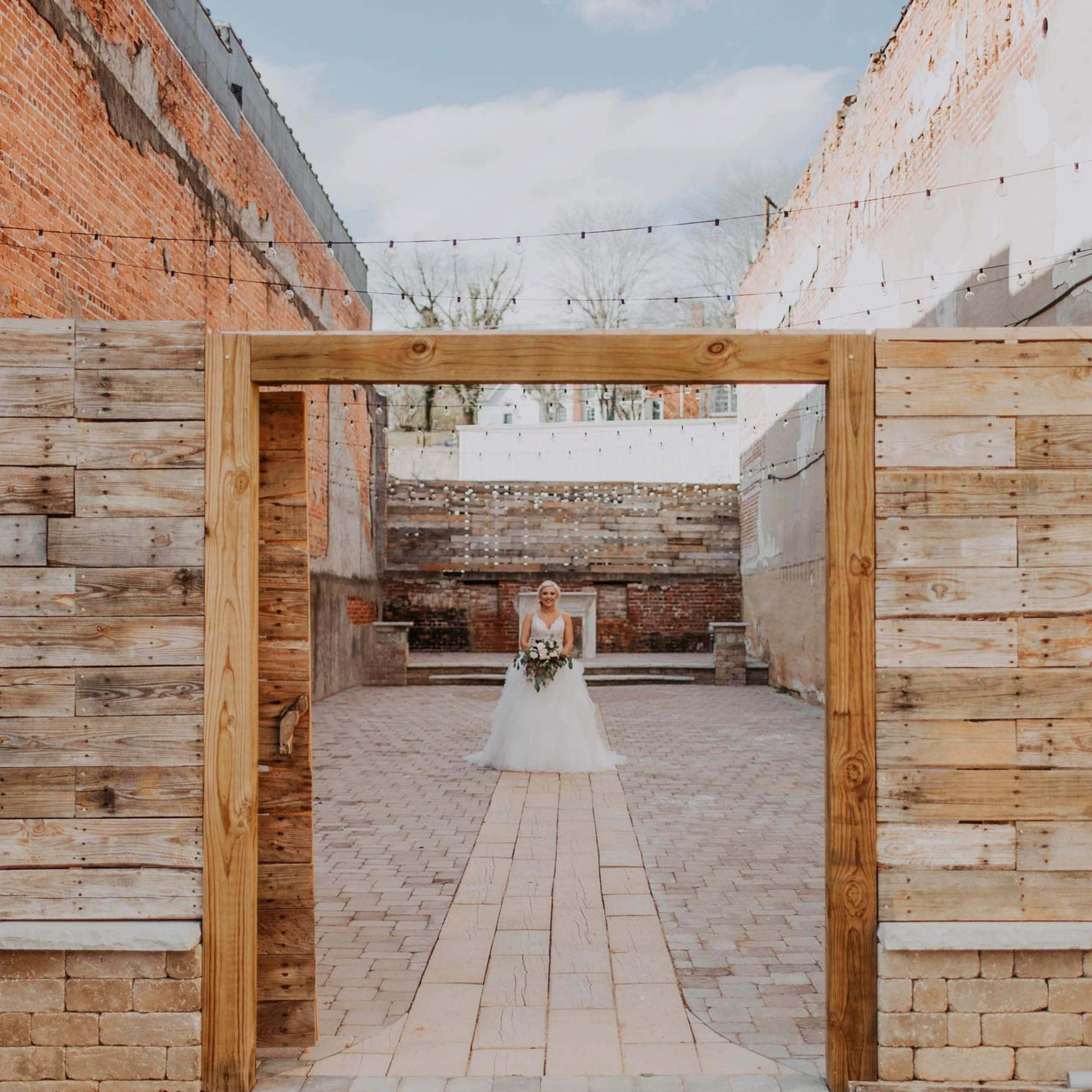 A bride in a white dress holds a bouquet as she stands in a courtyard surrounded by weathered brick walls and wooden structures.