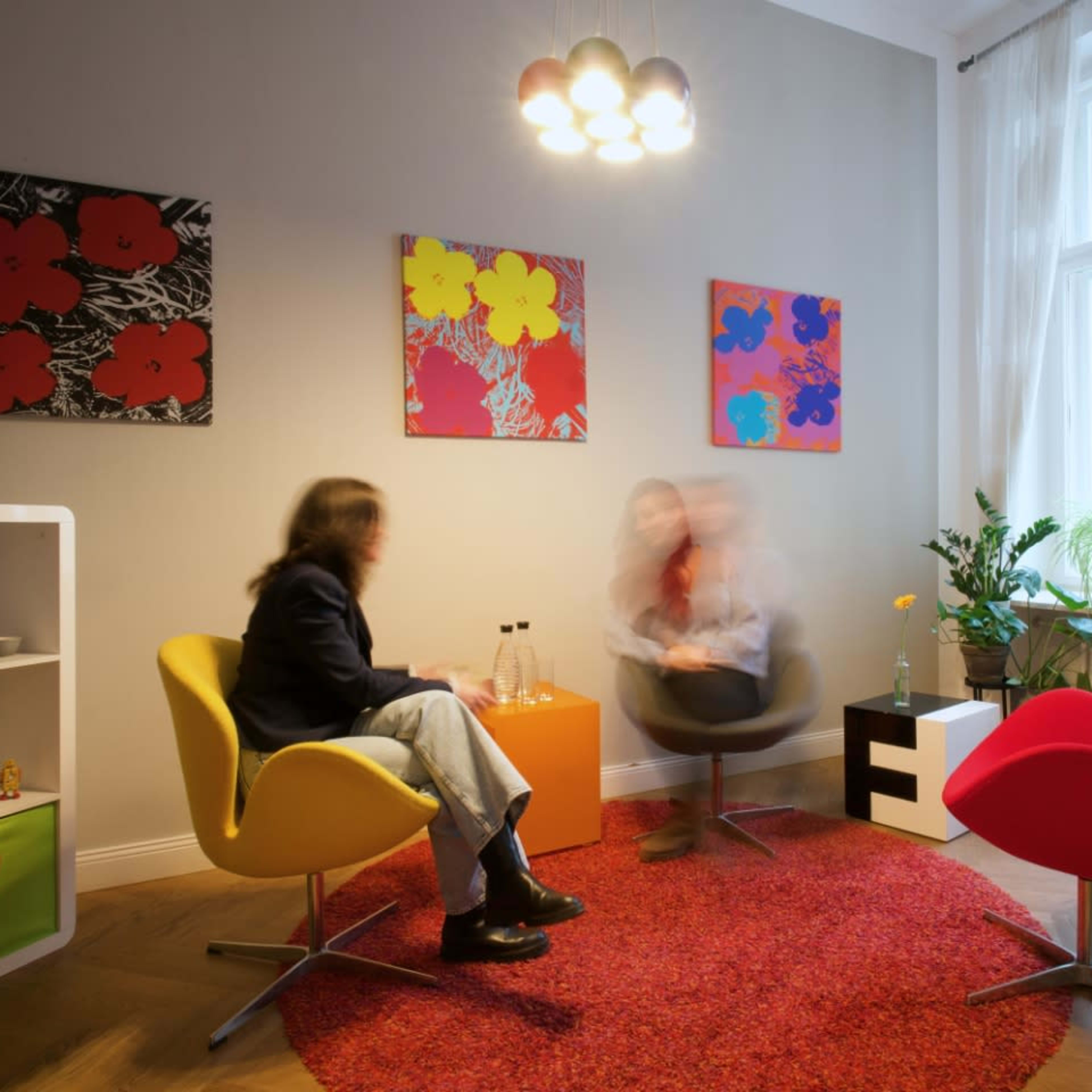 Two people are seated in colorful chairs in a modern room with abstract artwork on the walls and a round red rug on the floor.