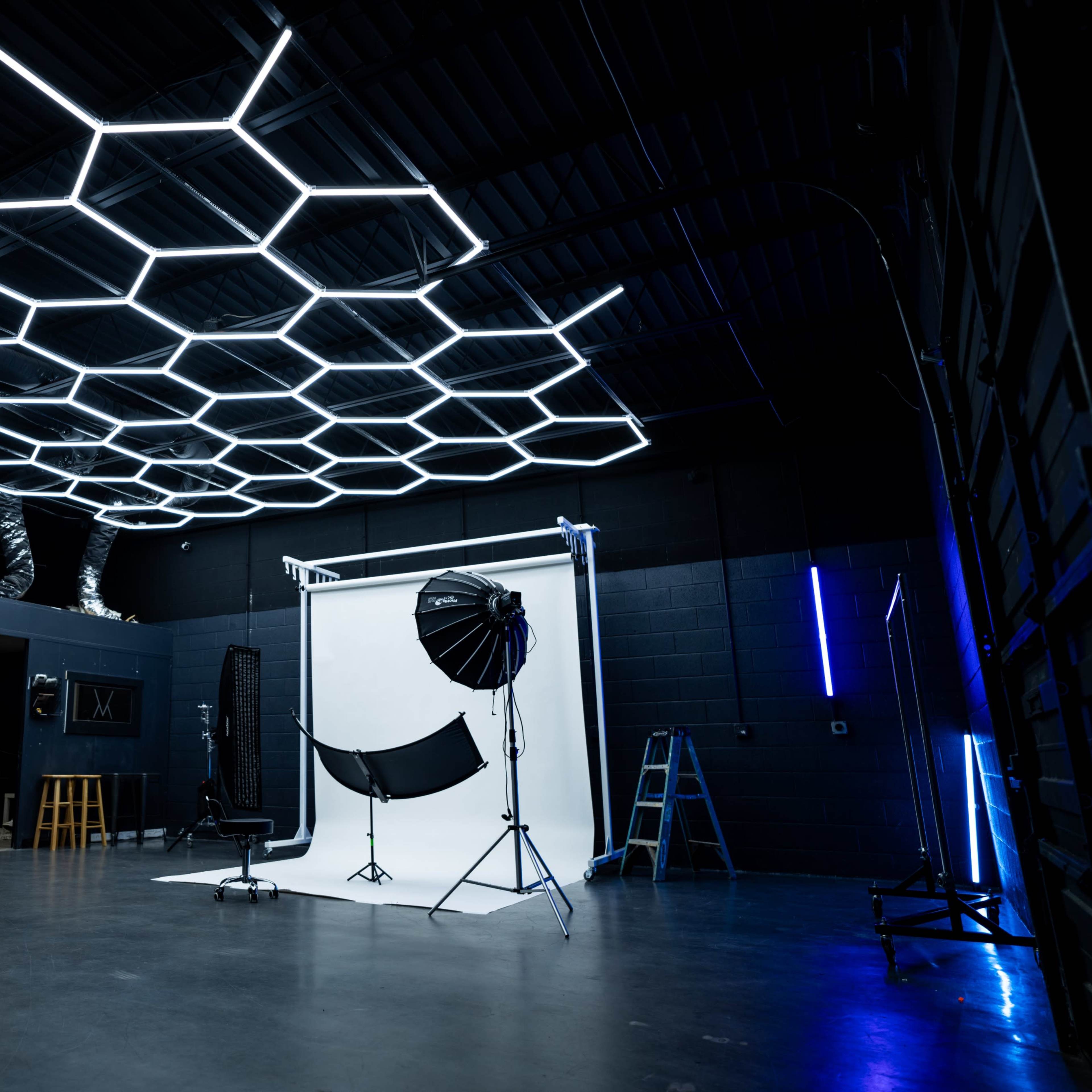 A photography studio with hexagonal LED lights overhead, a white backdrop setup, and various photography equipment arranged in the space.
