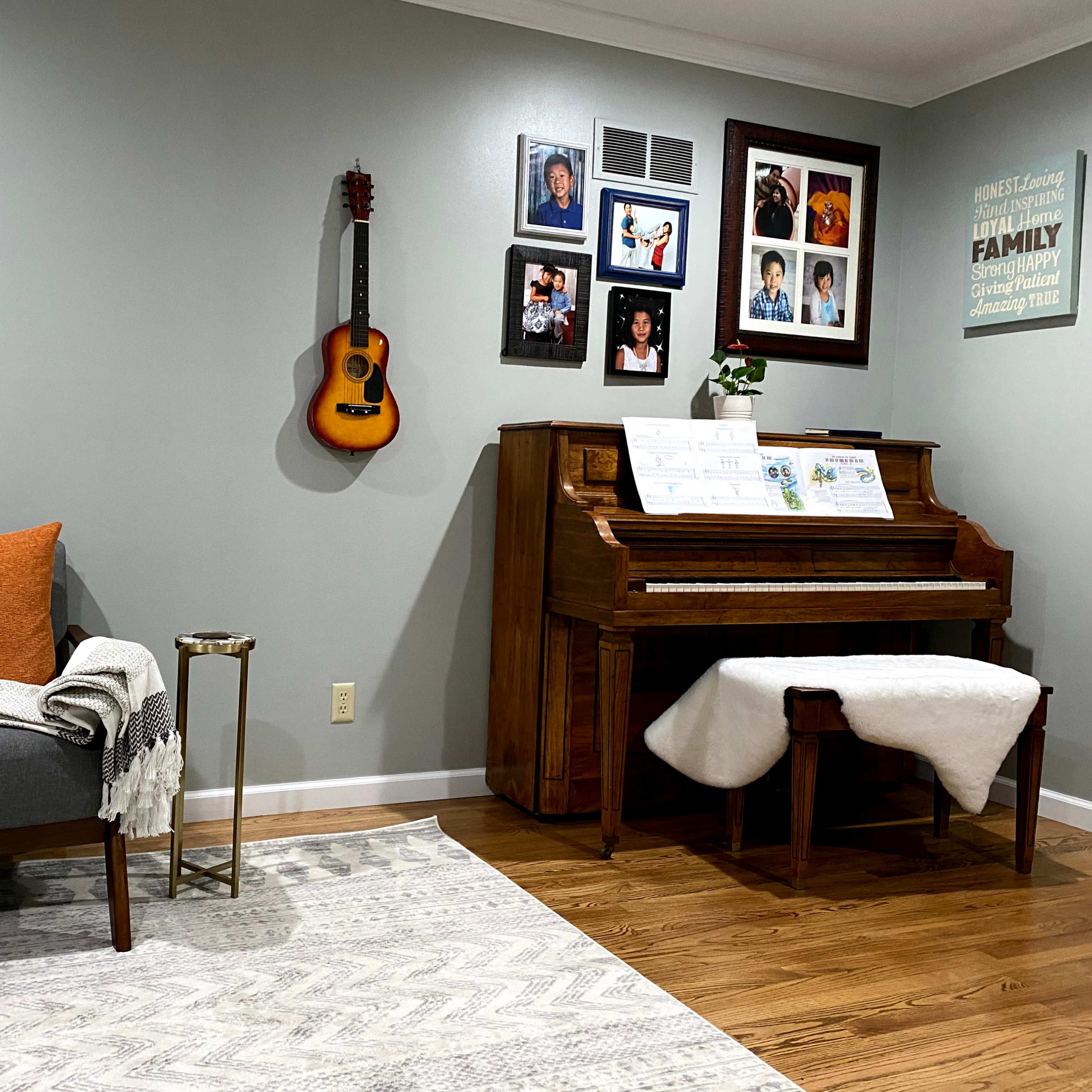 A cozy living room features a gray armchair with an orange pillow, an acoustic guitar and framed family photos on the wall, and a wooden piano with sheet music displayed.
