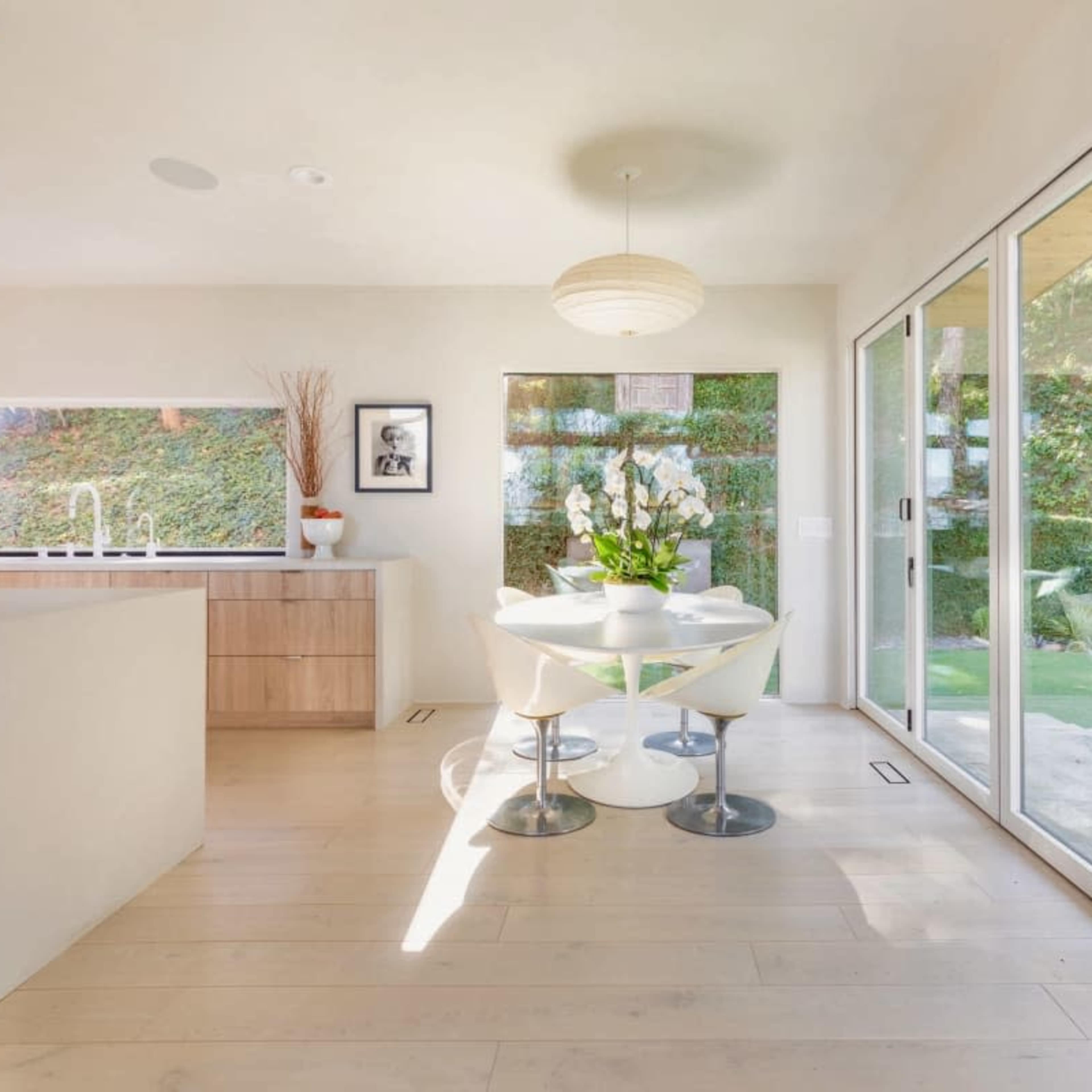 A bright kitchen and dining area features a round table with a floral centerpiece, large windows, and sliding glass doors leading to a garden.