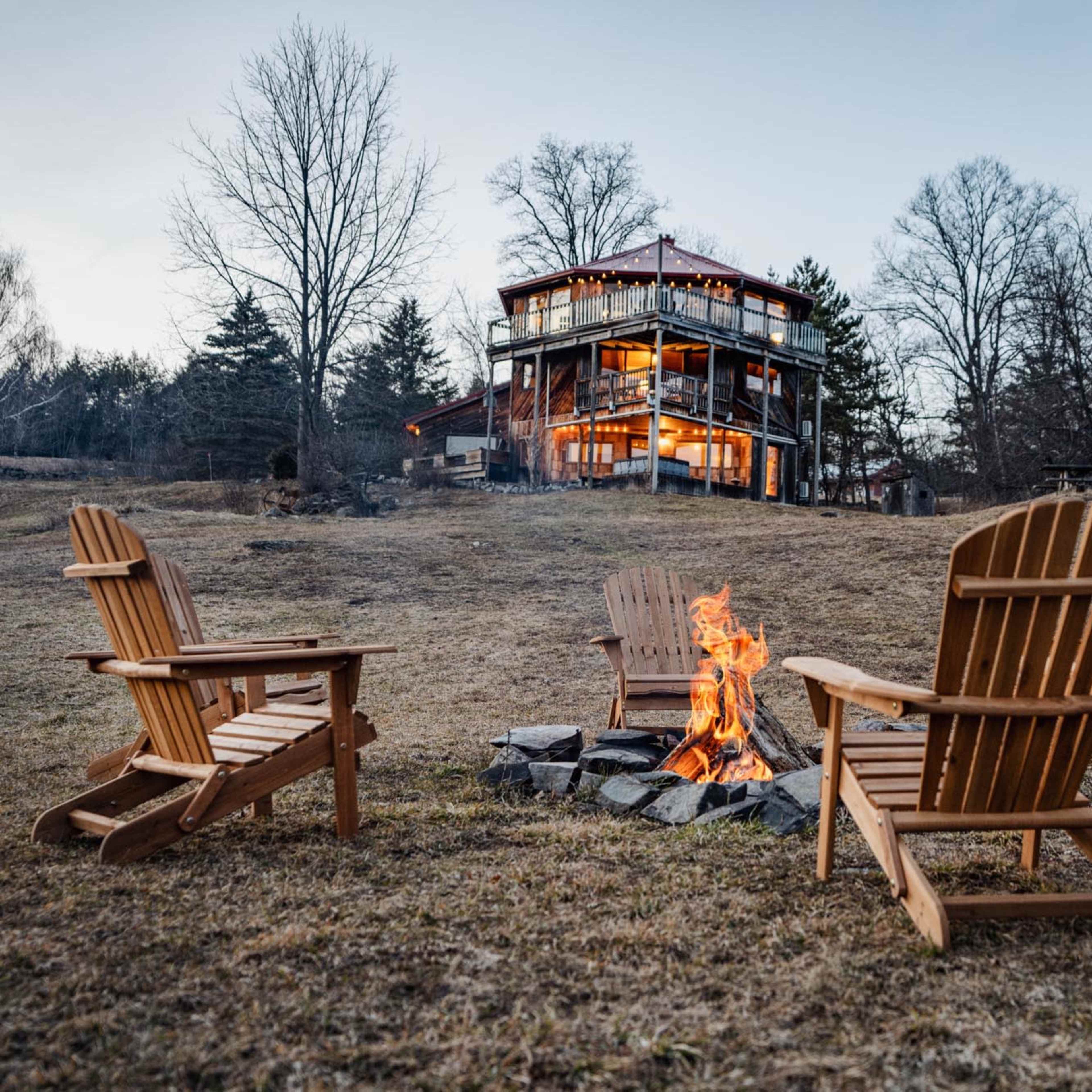 A circle of wooden chairs surrounds a fire pit with flames, while a multi-story building sits in the background against a twilight sky.