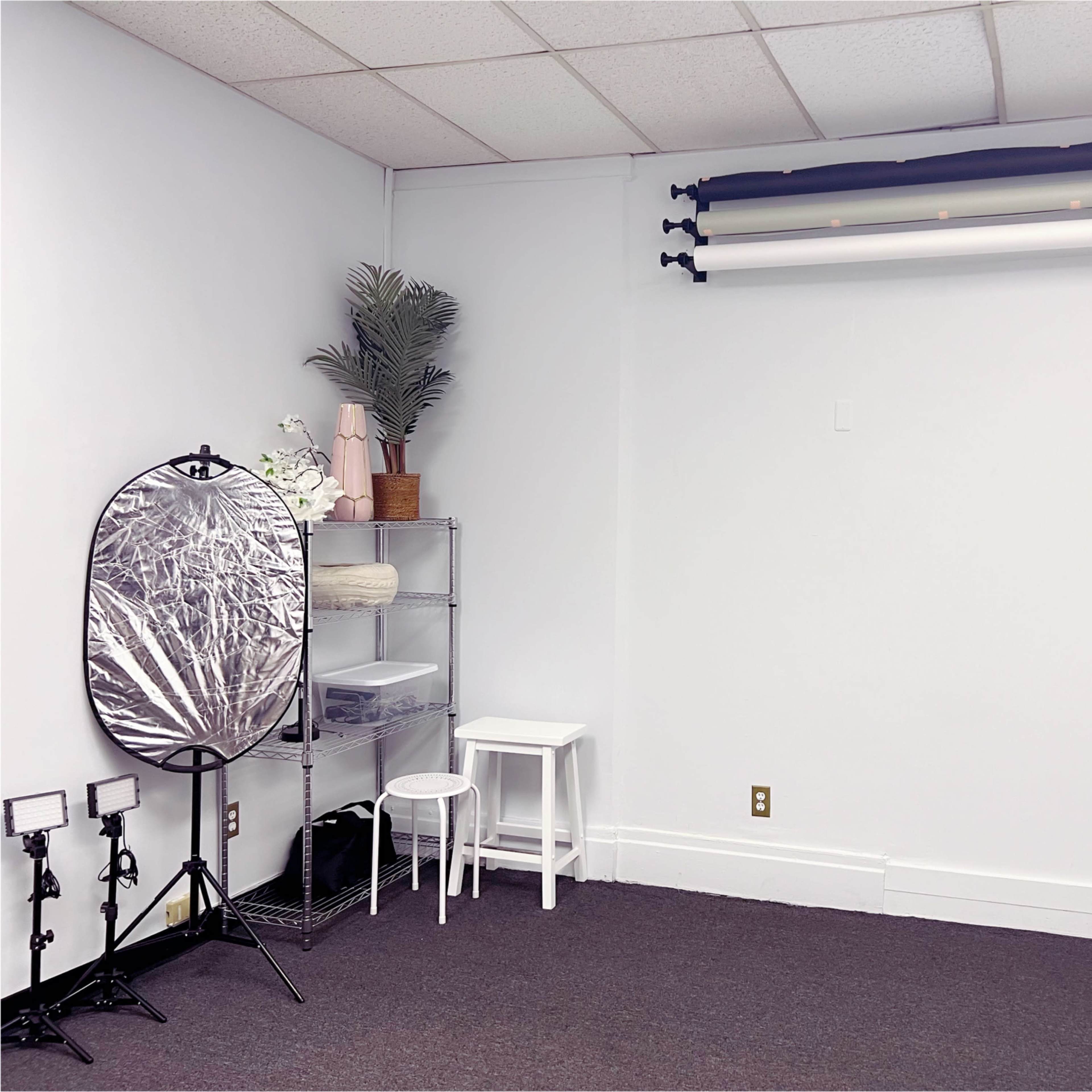 A minimalist studio space with a white wall, a rack of backdrop rolls, a reflective circular disc on a stand, a clear storage unit, a small white stool, and several light stands.