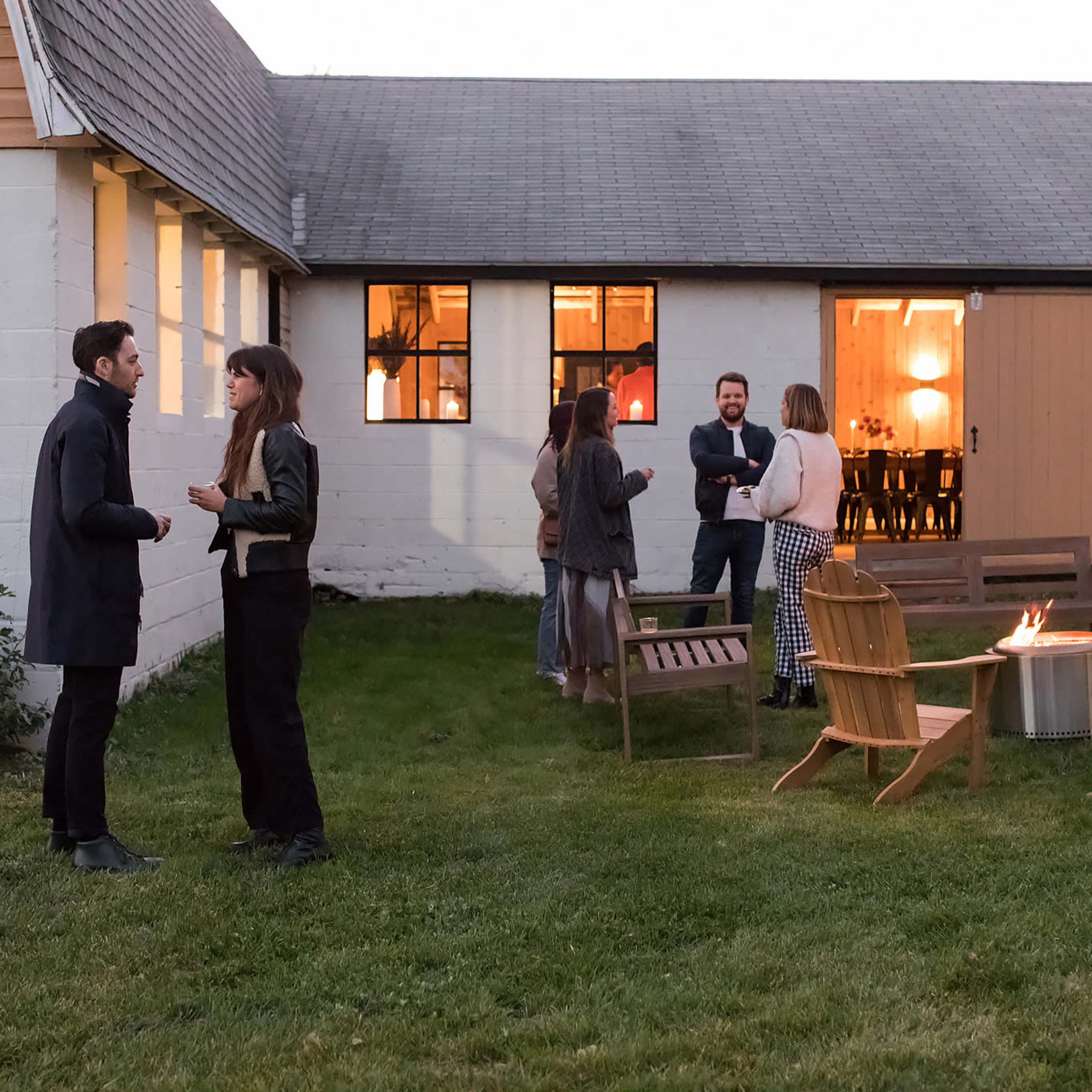 A gathering of five people in casual attire stands in a grassy area near a fire pit in front of a white building with large windows.