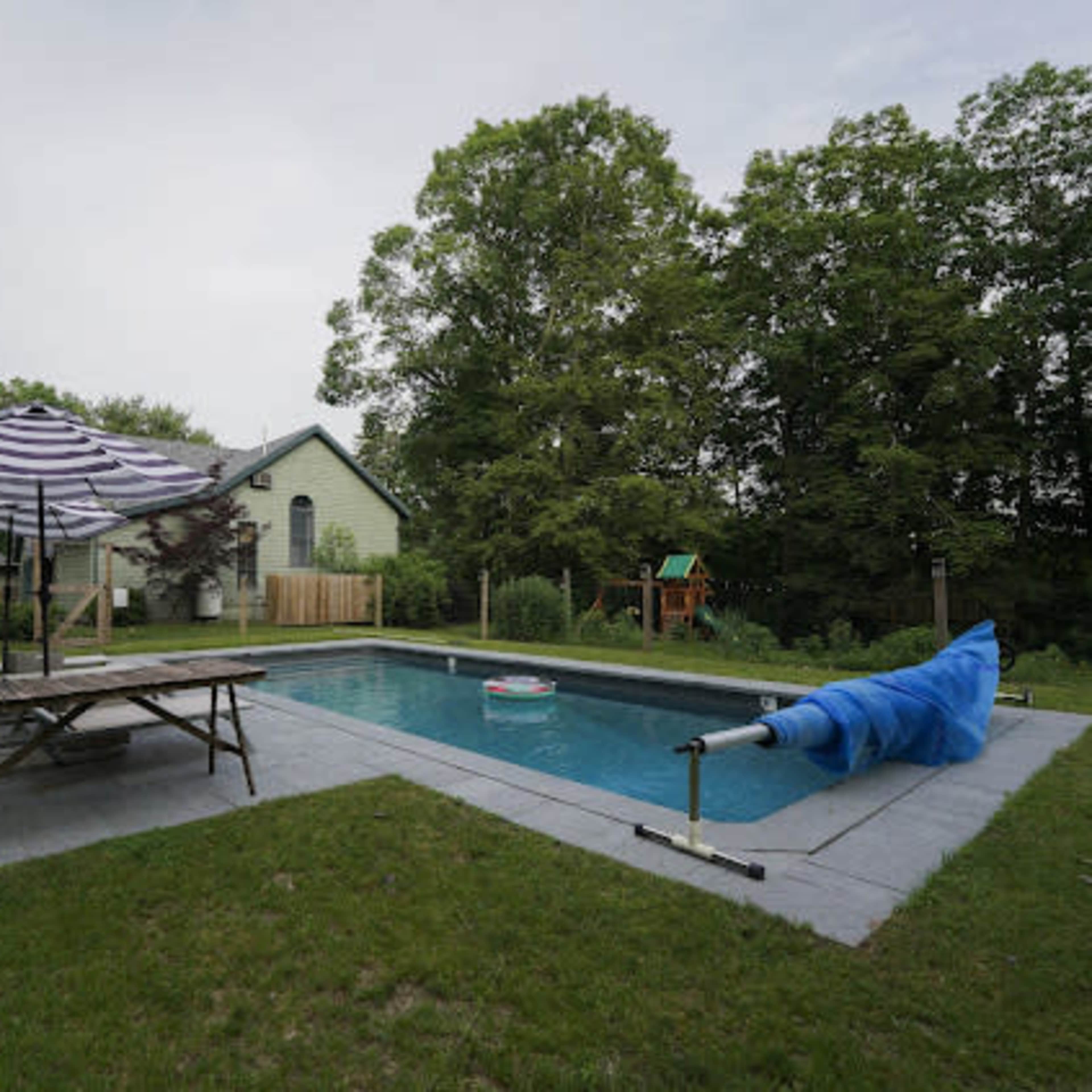 A backyard with a swimming pool surrounded by grass, a picnic table, an umbrella, and a house in the background.