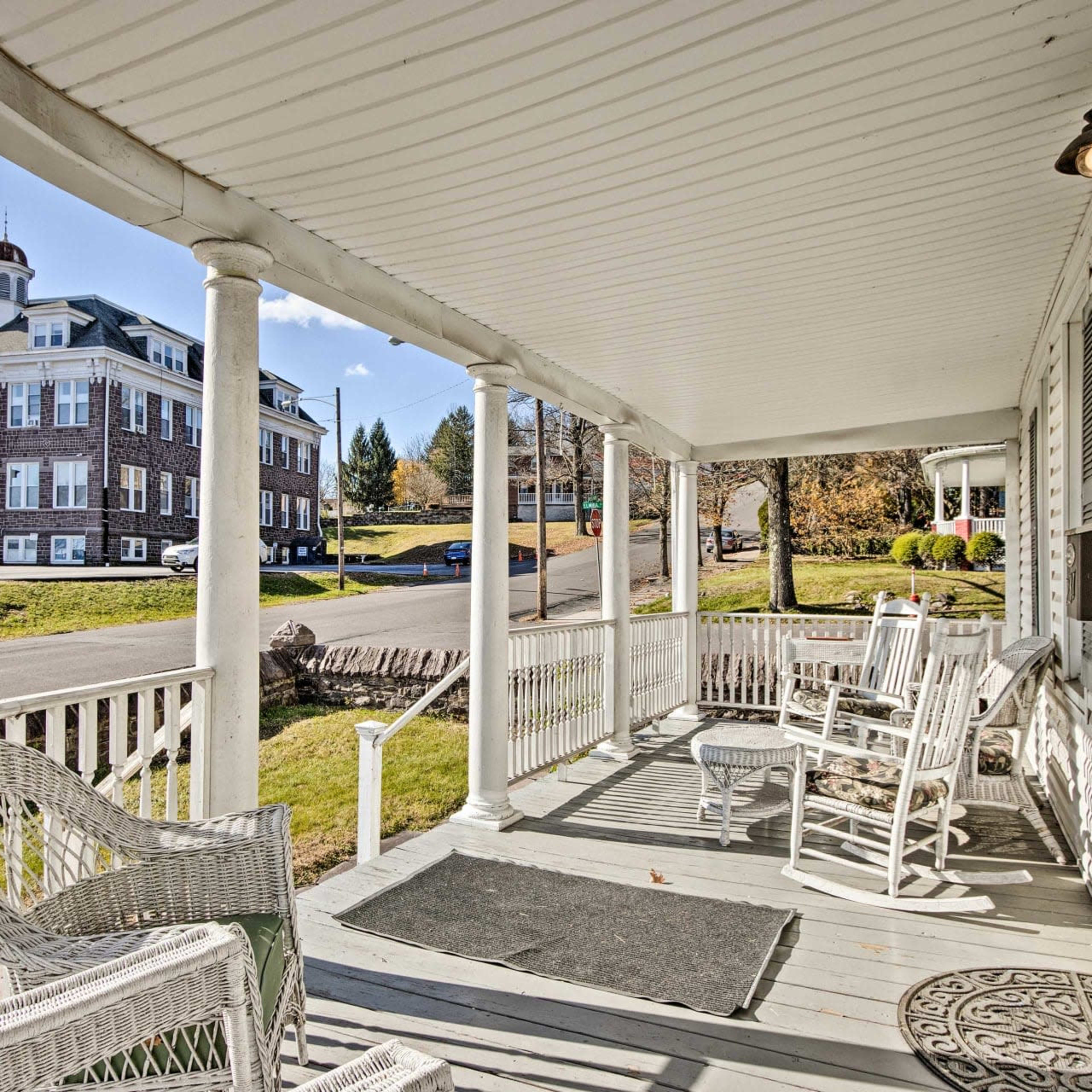 A porch with white rocking chairs, overlooking a street and a large building in the background.