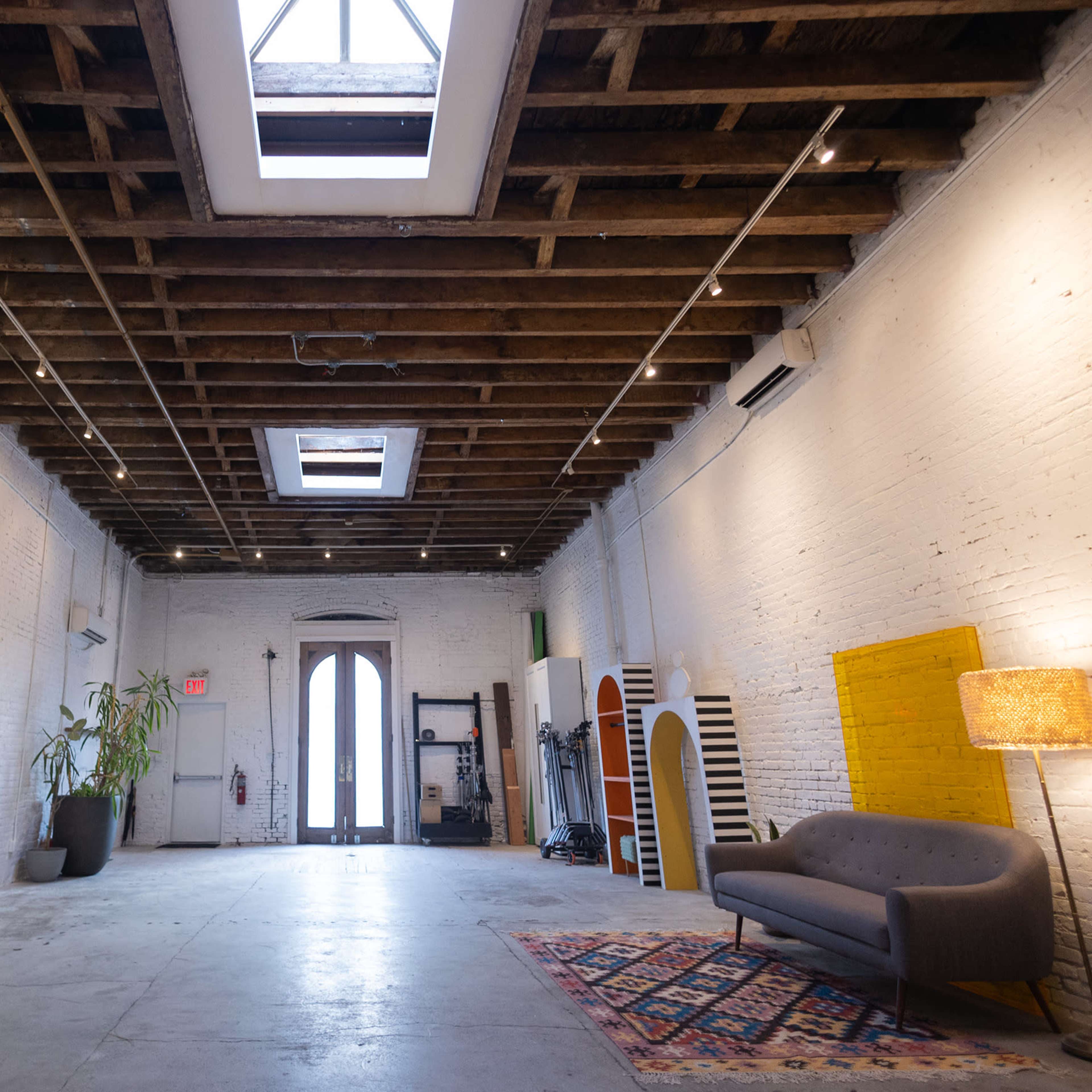 A spacious, minimalist room with exposed wooden beams, large windows, and a few pieces of furniture, including a gray sofa and a standing lamp.