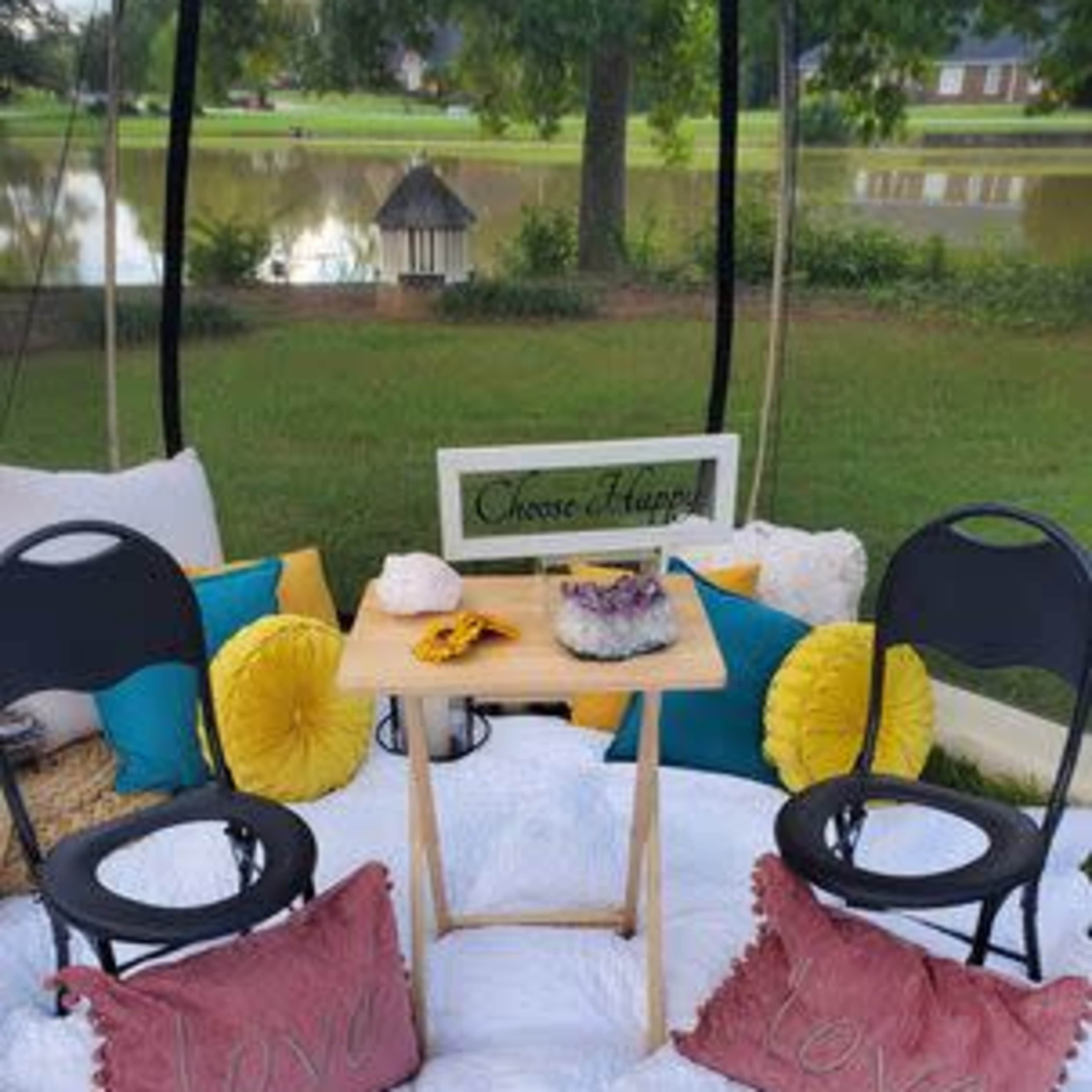 A cozy, outdoor seating area features two black chairs, a small table, pillows with the word "love," and colorful cushions arranged on a white blanket near a serene pond.