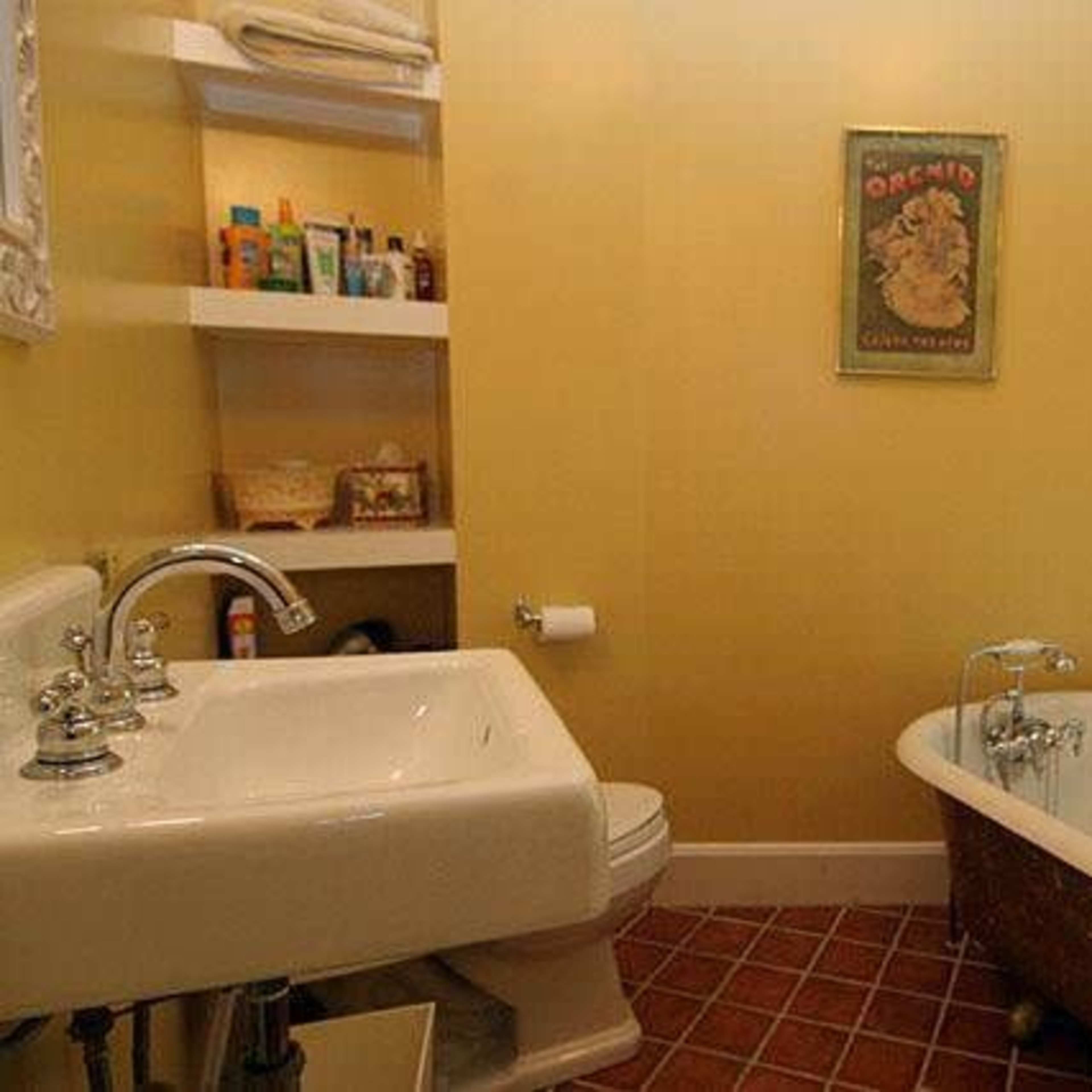 A small bathroom with a vintage clawfoot bathtub, a sink, and shelves containing various toiletries, all set against yellow walls and a window.