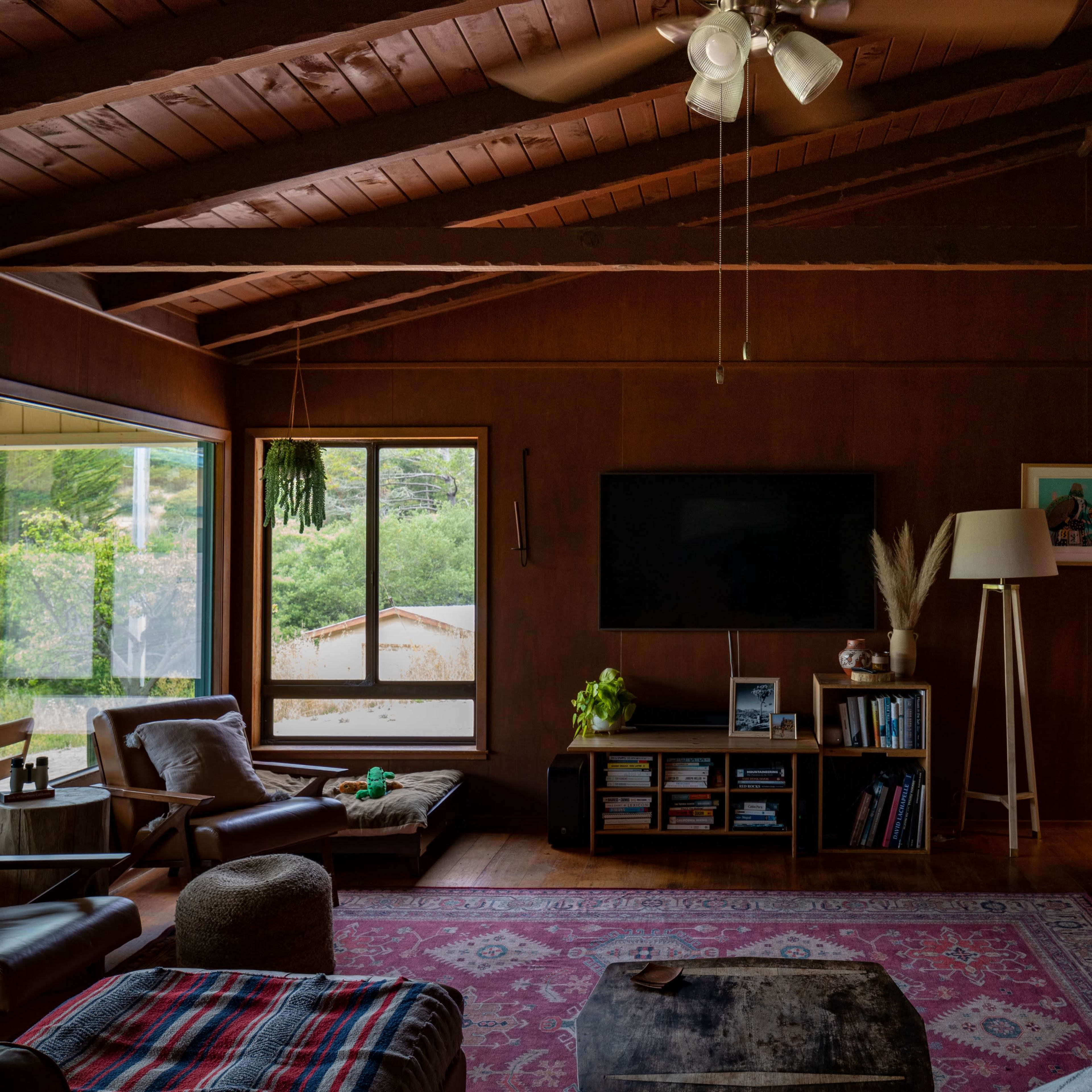 A living room with a stone fireplace, a television mounted on the wall, and large windows allowing natural light to enter.