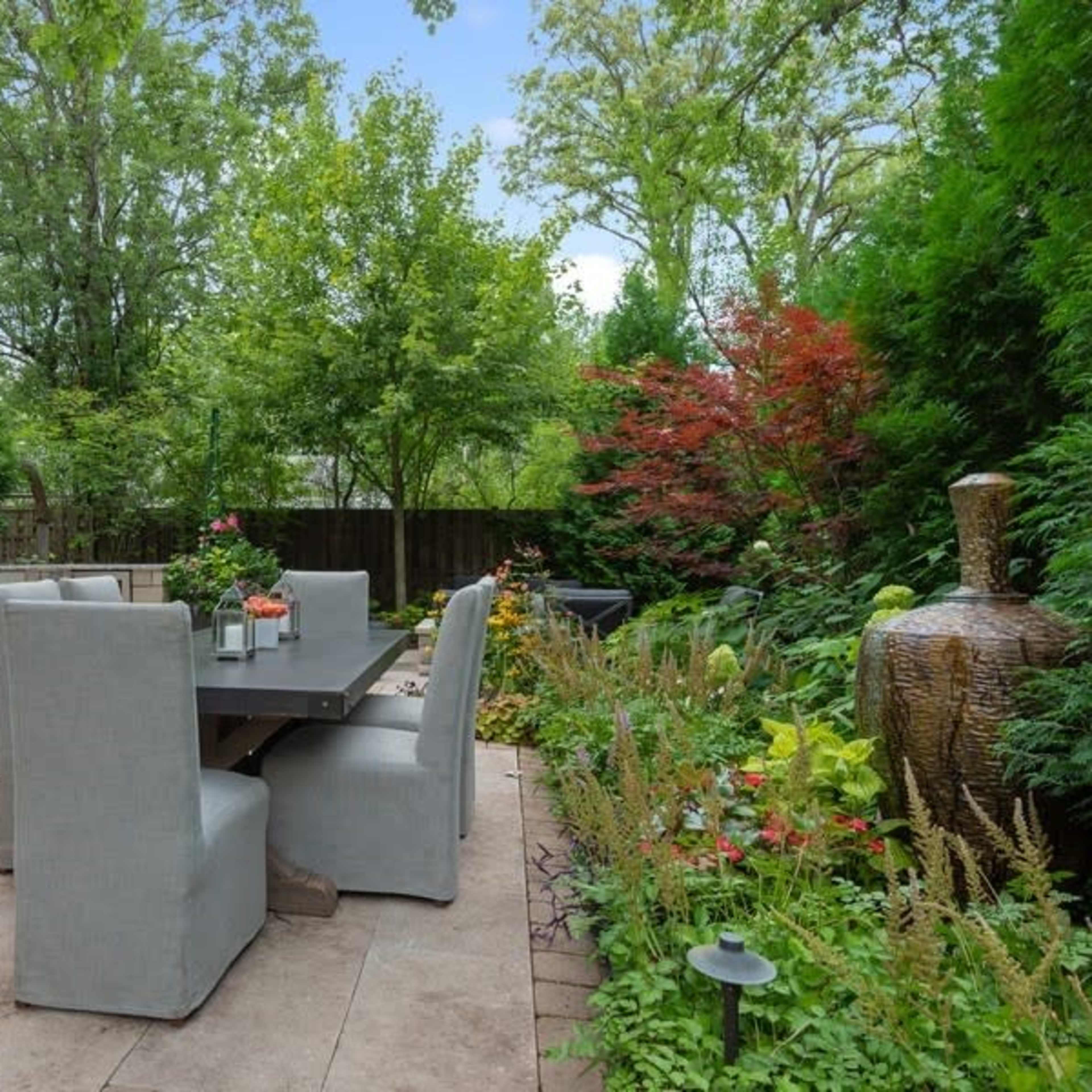 A patio area features a large dining table with gray chairs surrounded by greenery and landscaping elements.
