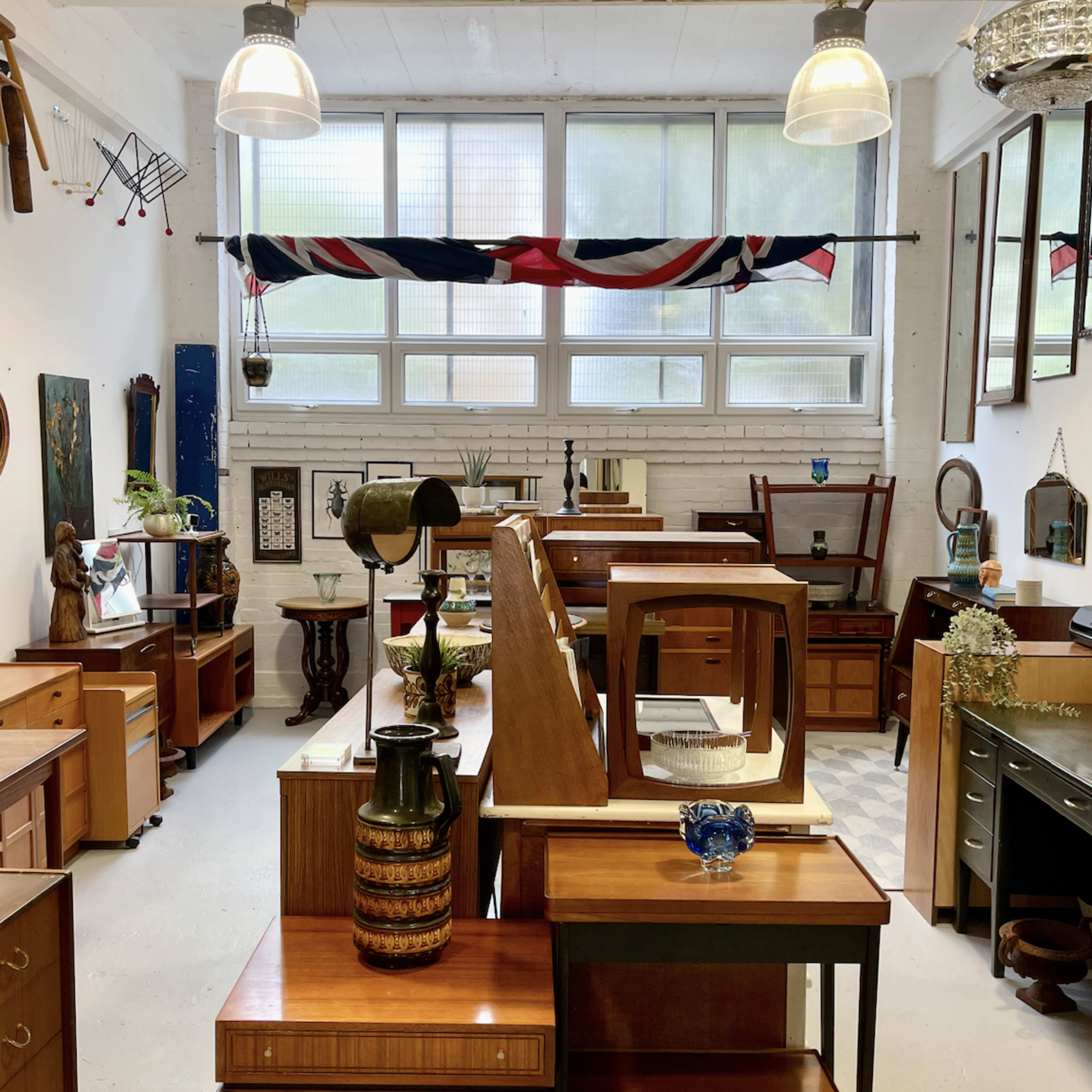 A spacious room filled with various wooden furniture pieces, including tables, cabinets, and decorative items, under large windows with a British flag hanging across the top.