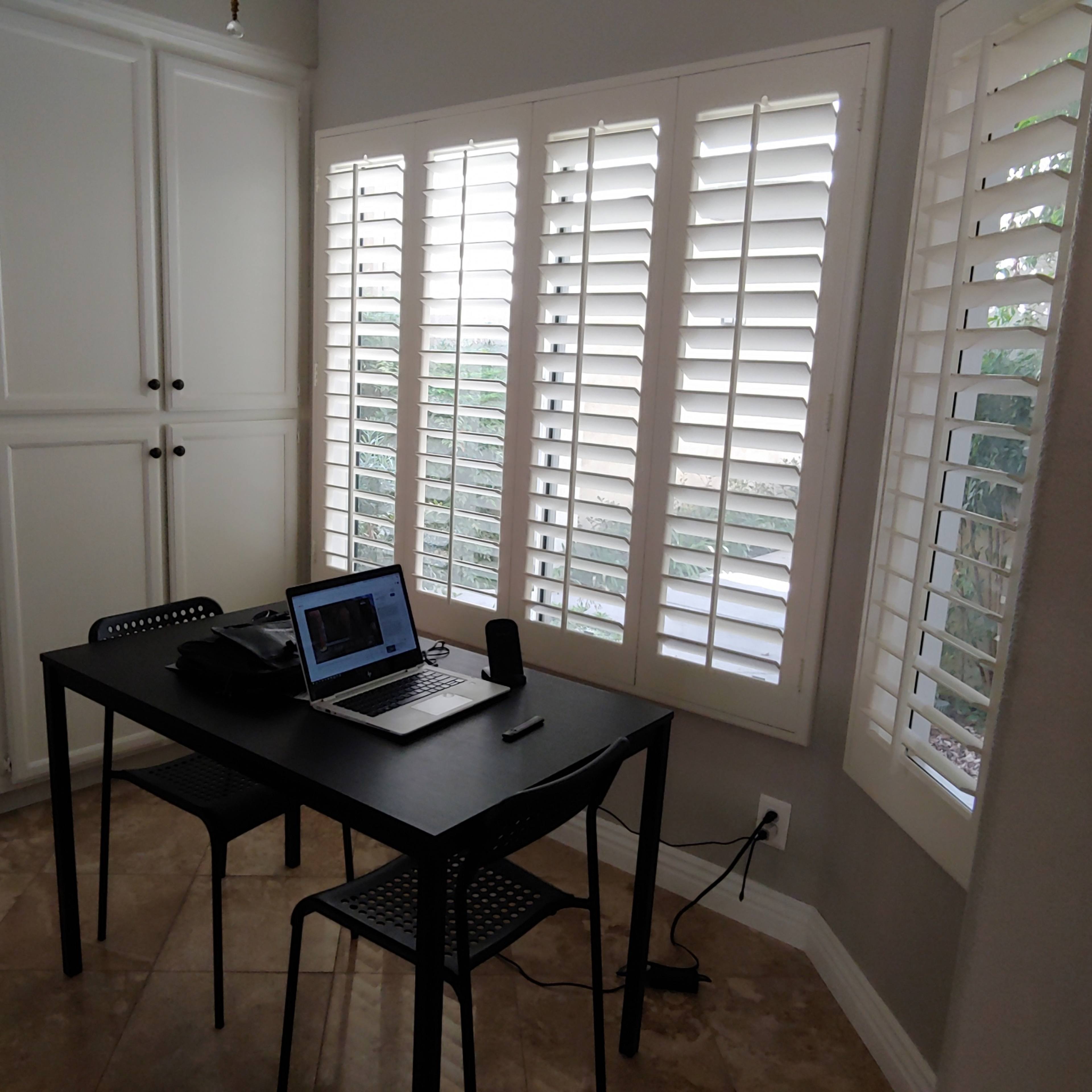 A small table with a laptop and a speaker is positioned in a well-lit room featuring plantation shutters and tiled flooring.