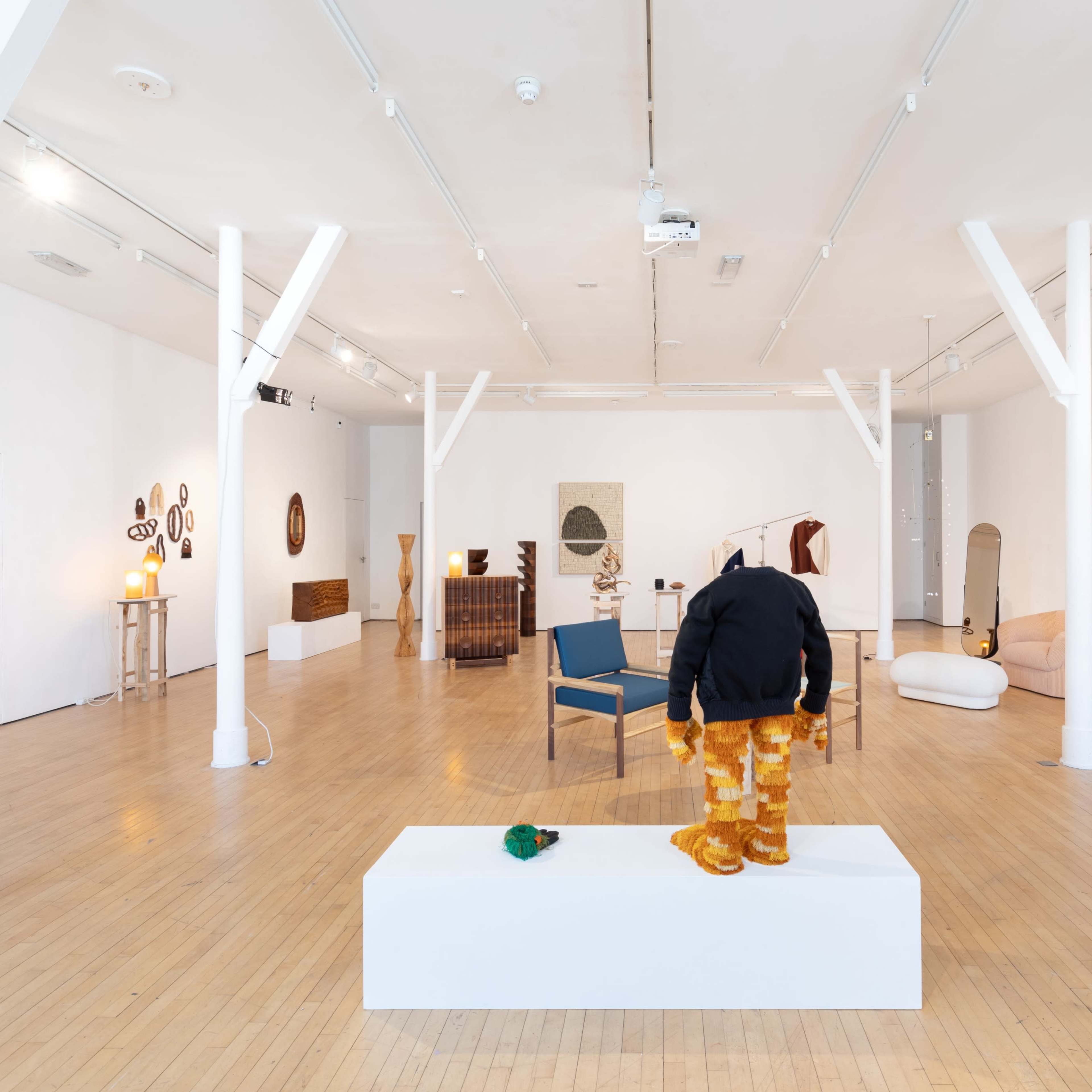 A contemporary art gallery featuring various sculptures and furniture pieces arranged in a spacious, well-lit room with white walls and wooden flooring.