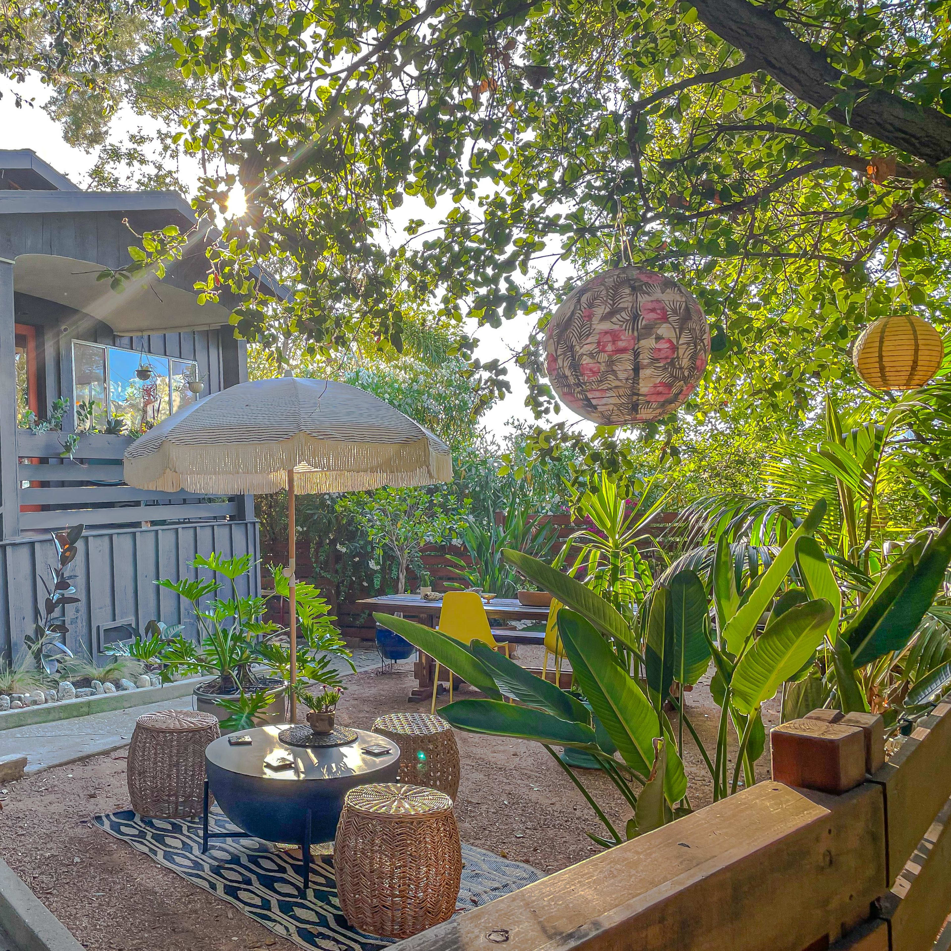A garden area features a patio with an umbrella, decorative lanterns, and a seating arrangement surrounded by plants and a house in the background.
