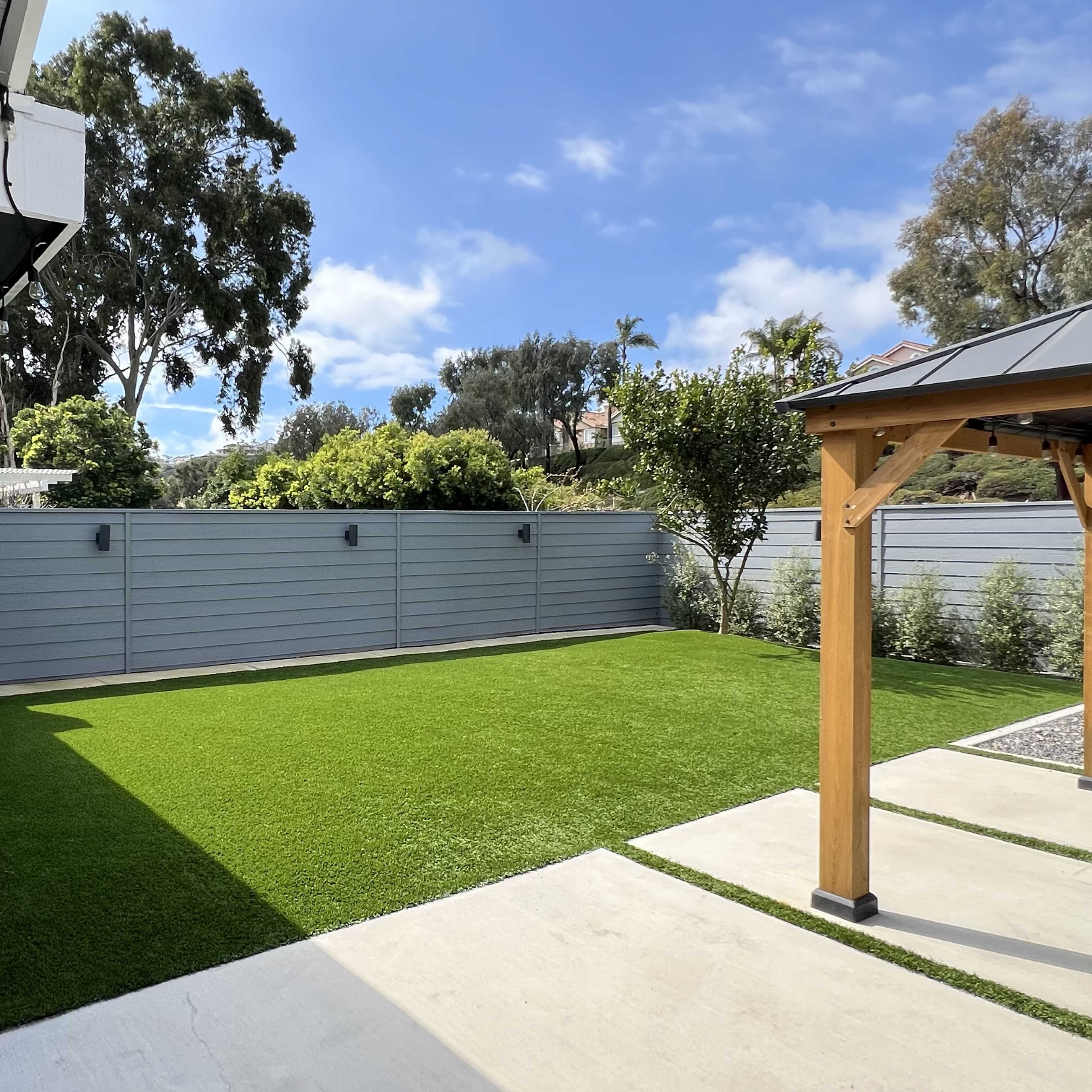 A landscaped backyard with artificial grass, a wooden pergola, and a gray fence under a partly cloudy sky.