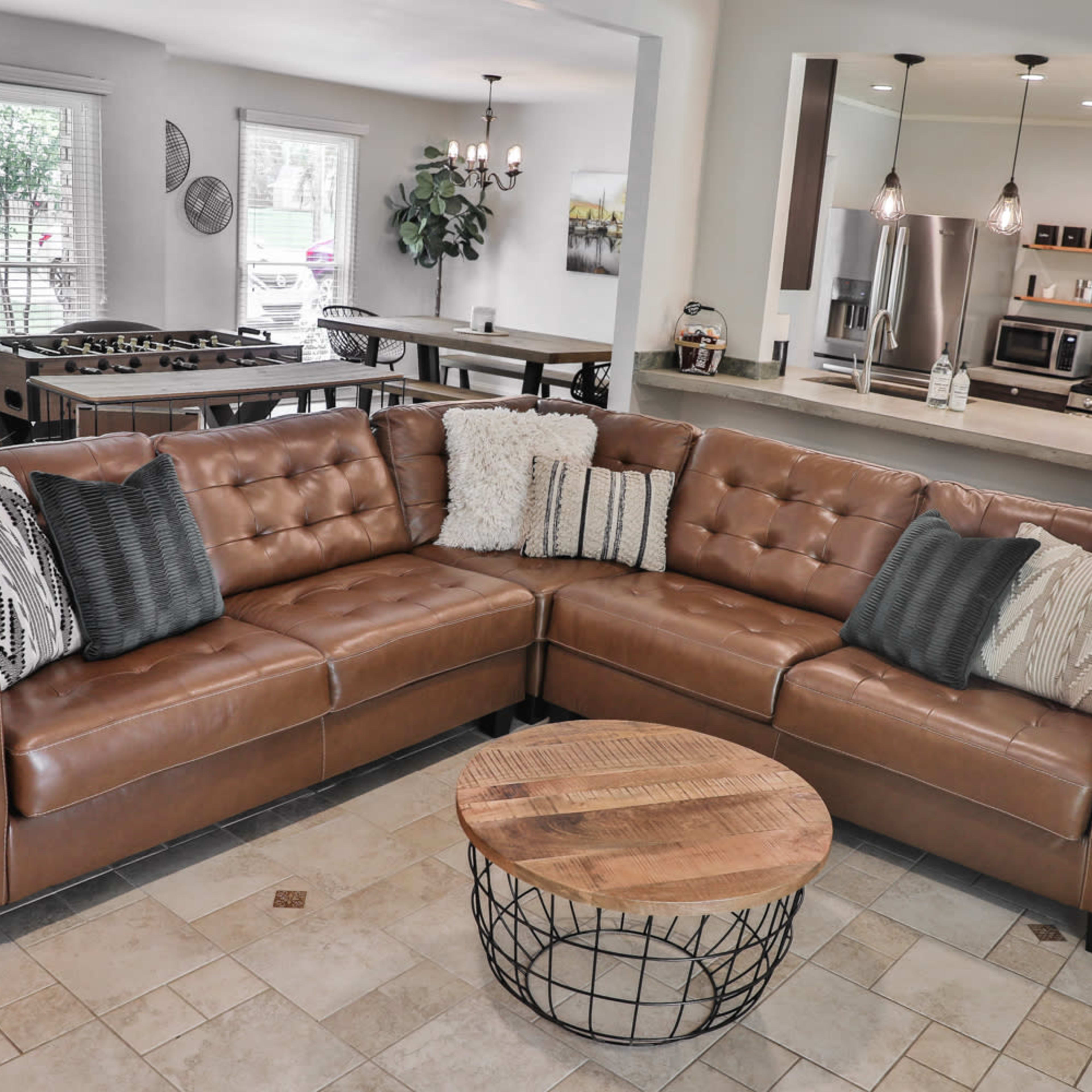 A modern living area featuring a brown leather sectional sofa with decorative pillows, a circular coffee table, and a view into a kitchen and dining space in the background.