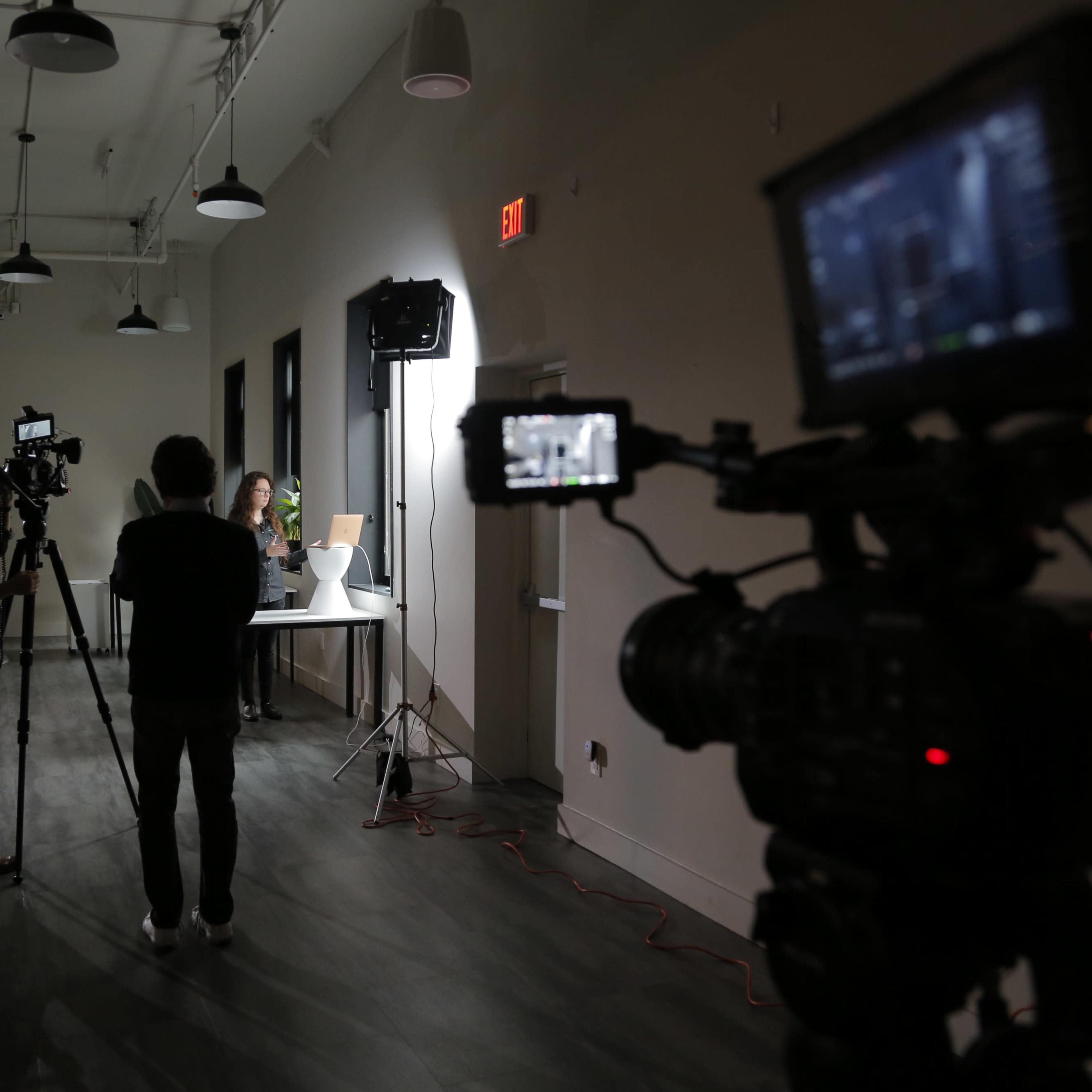 A filming setup in a dimly lit room features two cameras on tripods facing a person at a table with lighting equipment in the background.