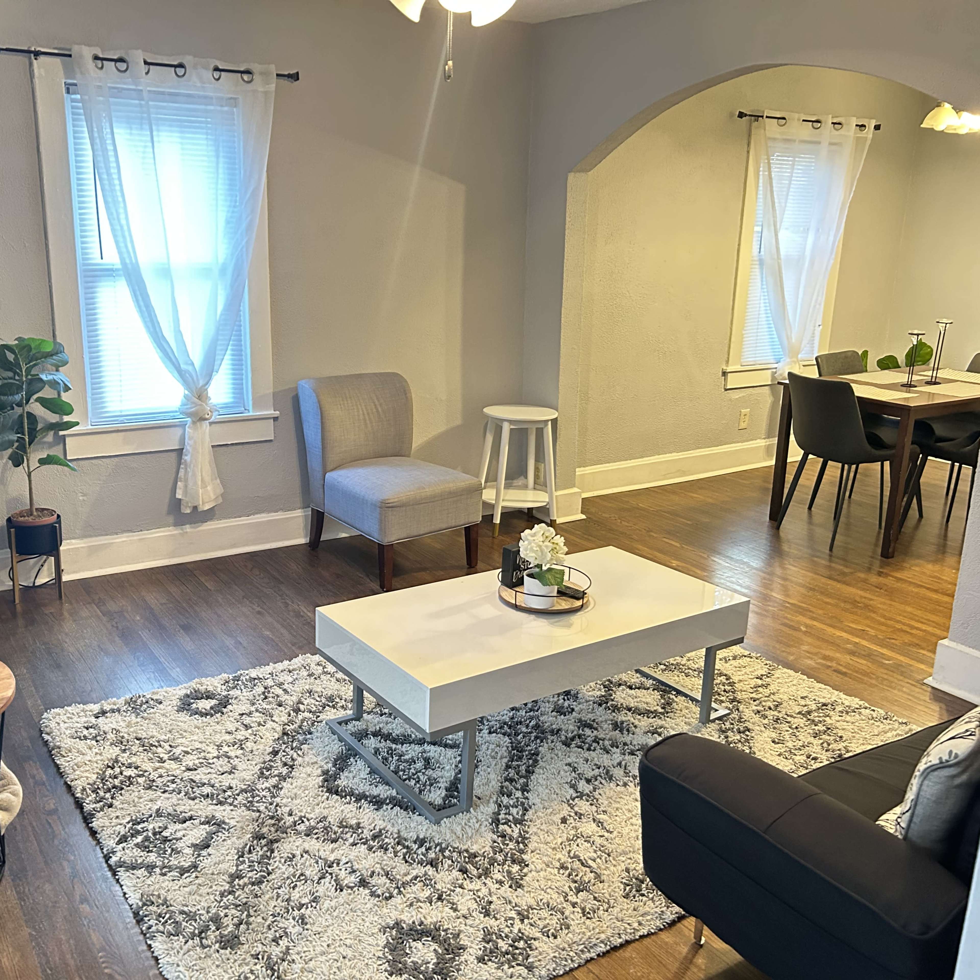 A cozy living room with a gray color scheme, featuring a coffee table, chairs, and a plant, connected to a dining area in the background.