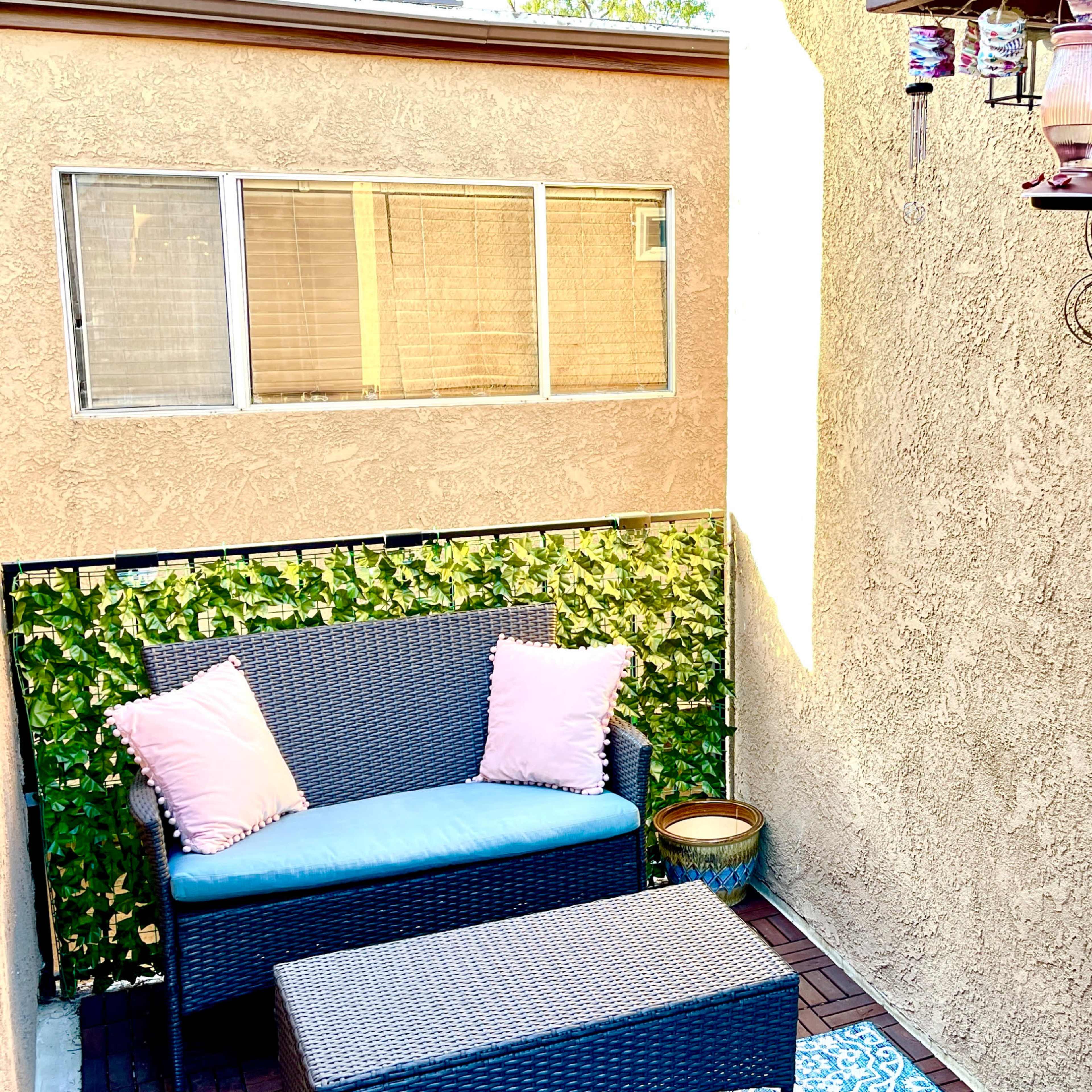 A small outdoor patio with a wicker loveseat, a matching coffee table, and decorative plants along the walls.