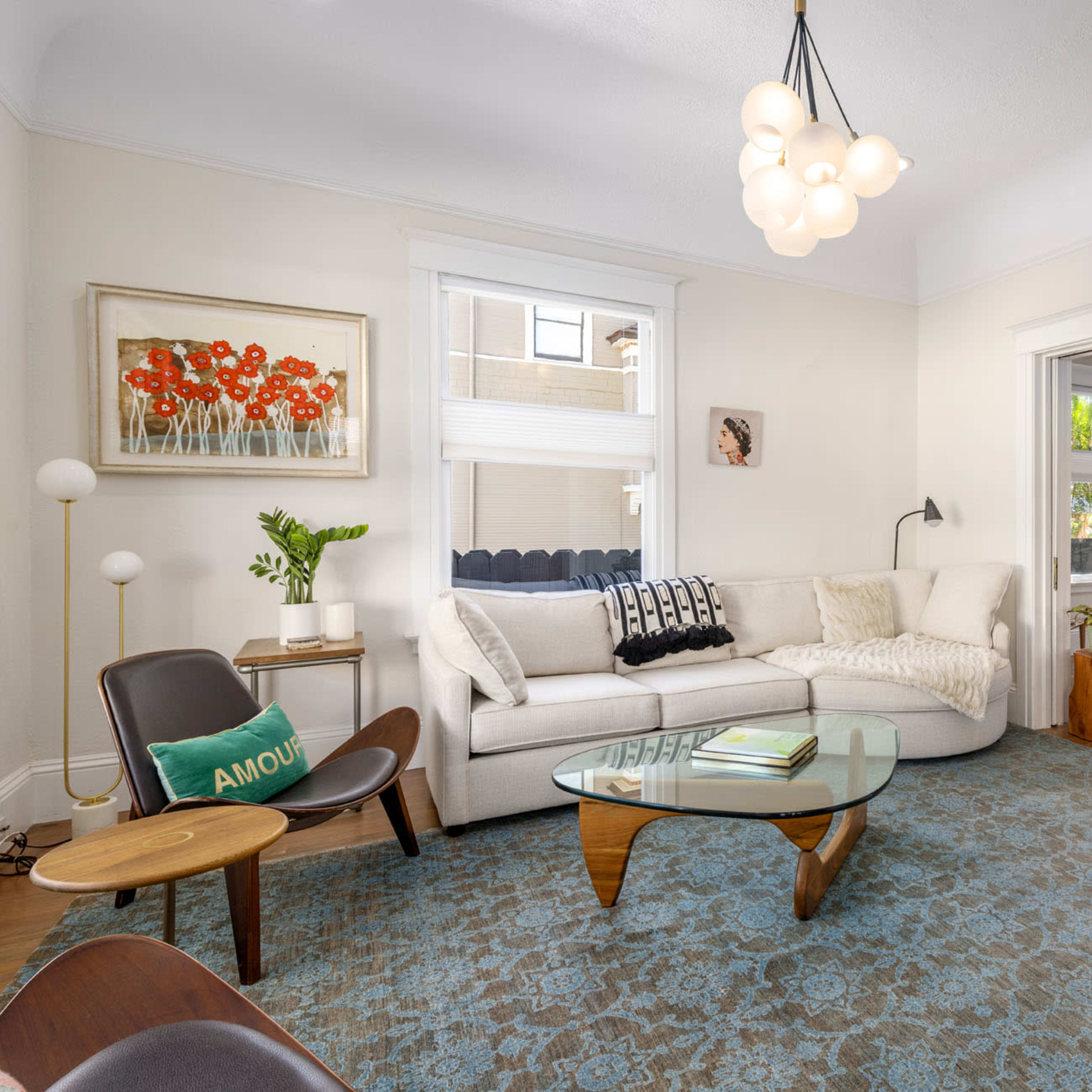 A well-lit living room featuring a white sectional sofa, a glass coffee table, and decorative plants, with an adjoining room visible in the background.