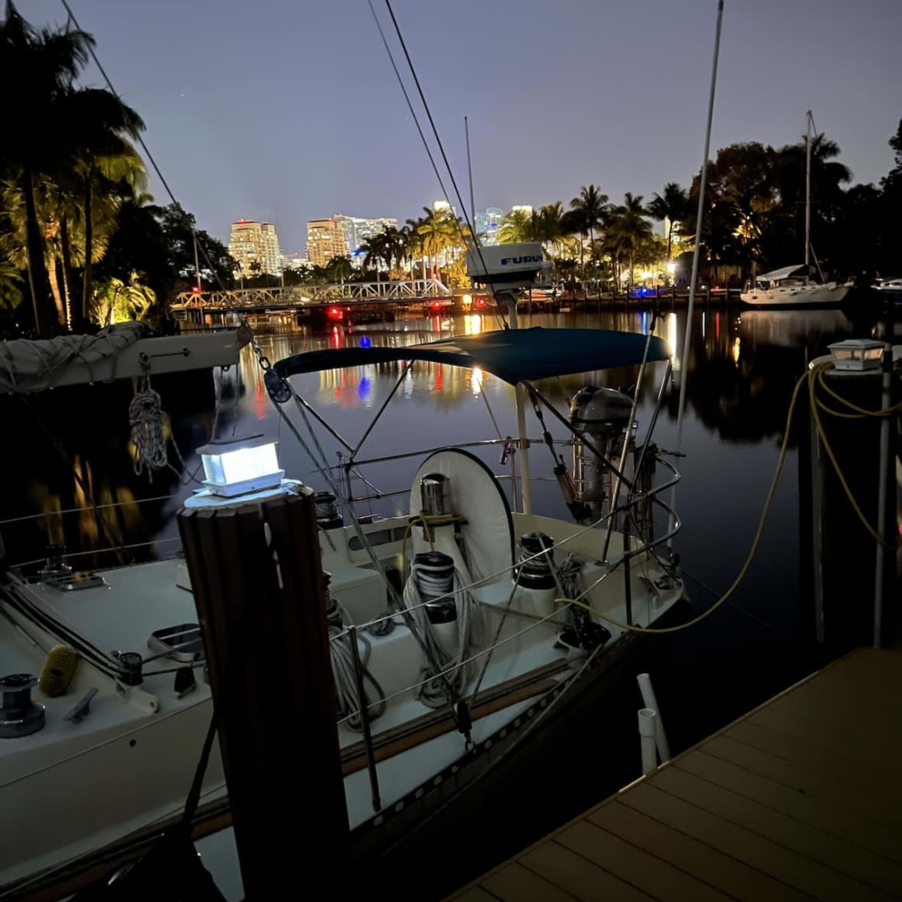 A sailboat is moored at a marina with city lights reflecting on the water at night.