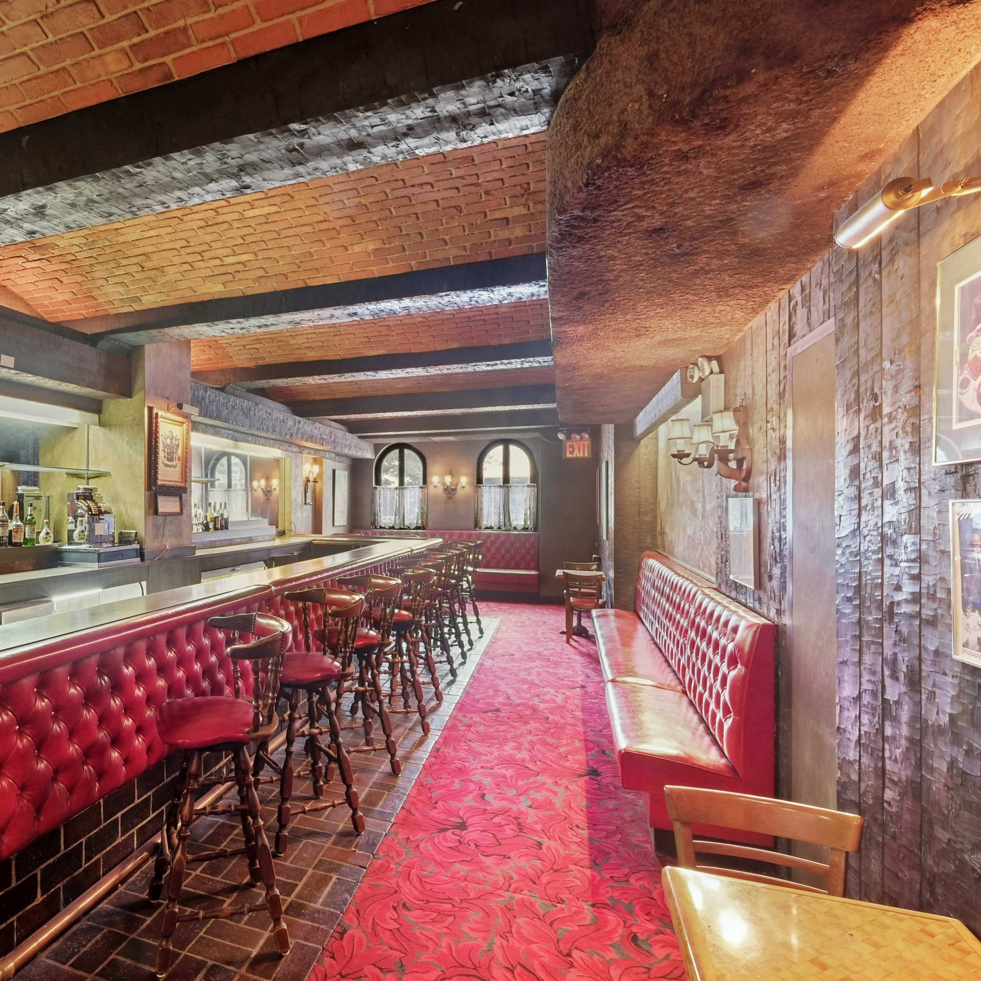 A dimly lit bar with red upholstered seating, a long marble-topped counter, and shelves lined with bottles in the background.