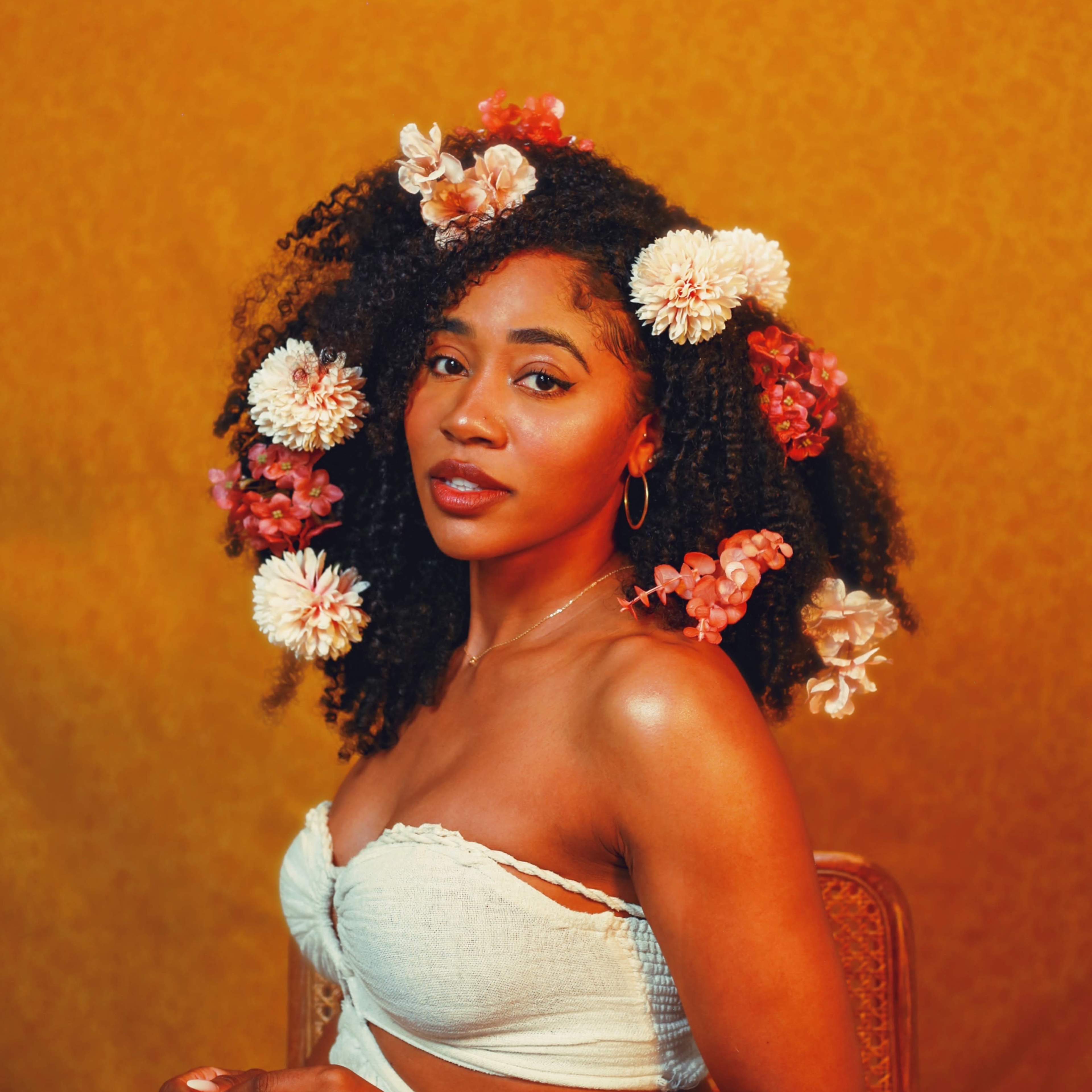 A woman with curly hair adorned with flowers poses confidently against a textured yellow background.