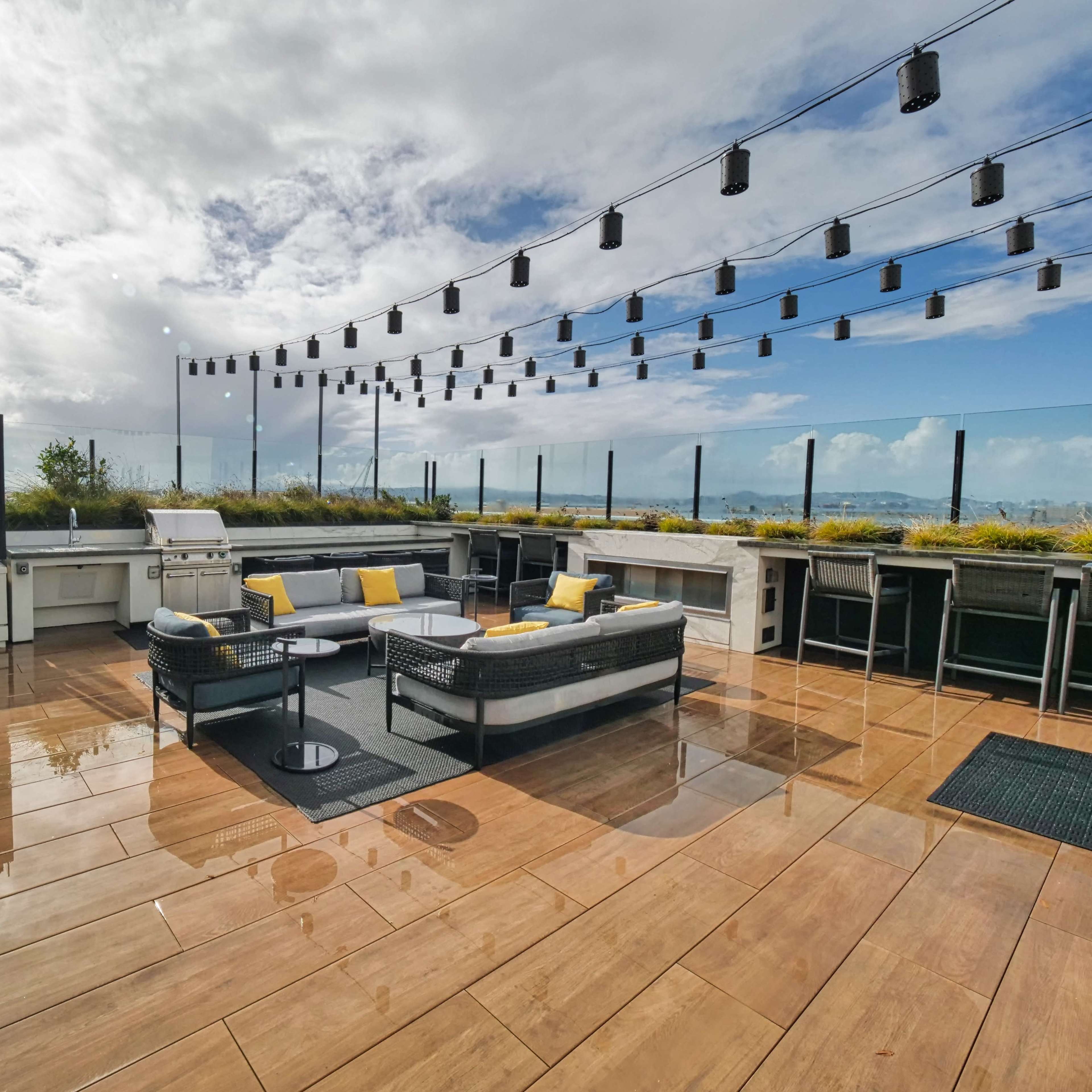 A rooftop terrace with modern furniture, outdoor kitchen facilities, and overhead string lights against a partly cloudy sky.