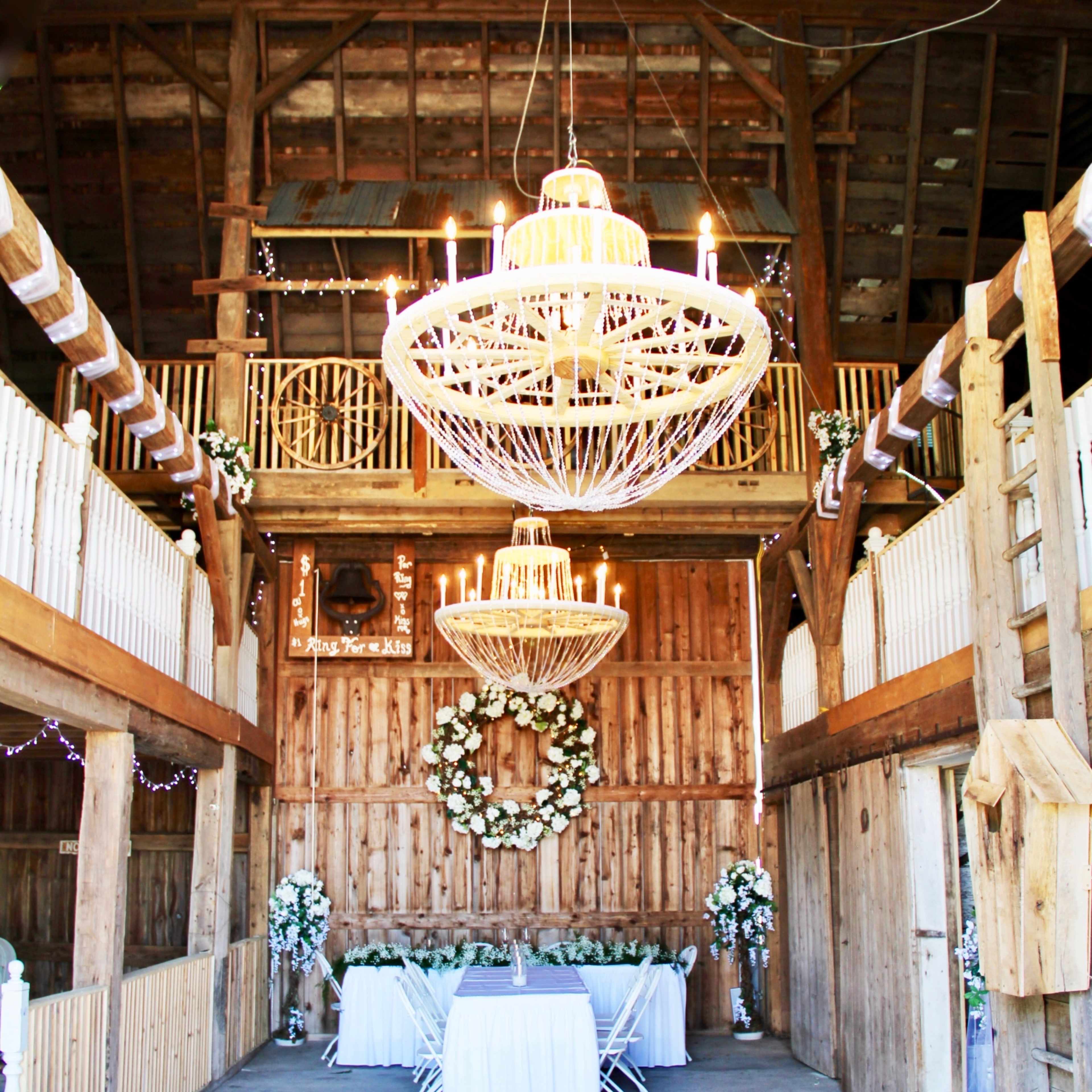 The interior of a barn features two large chandeliers, wooden walls, and a decorated table set for a wedding reception.