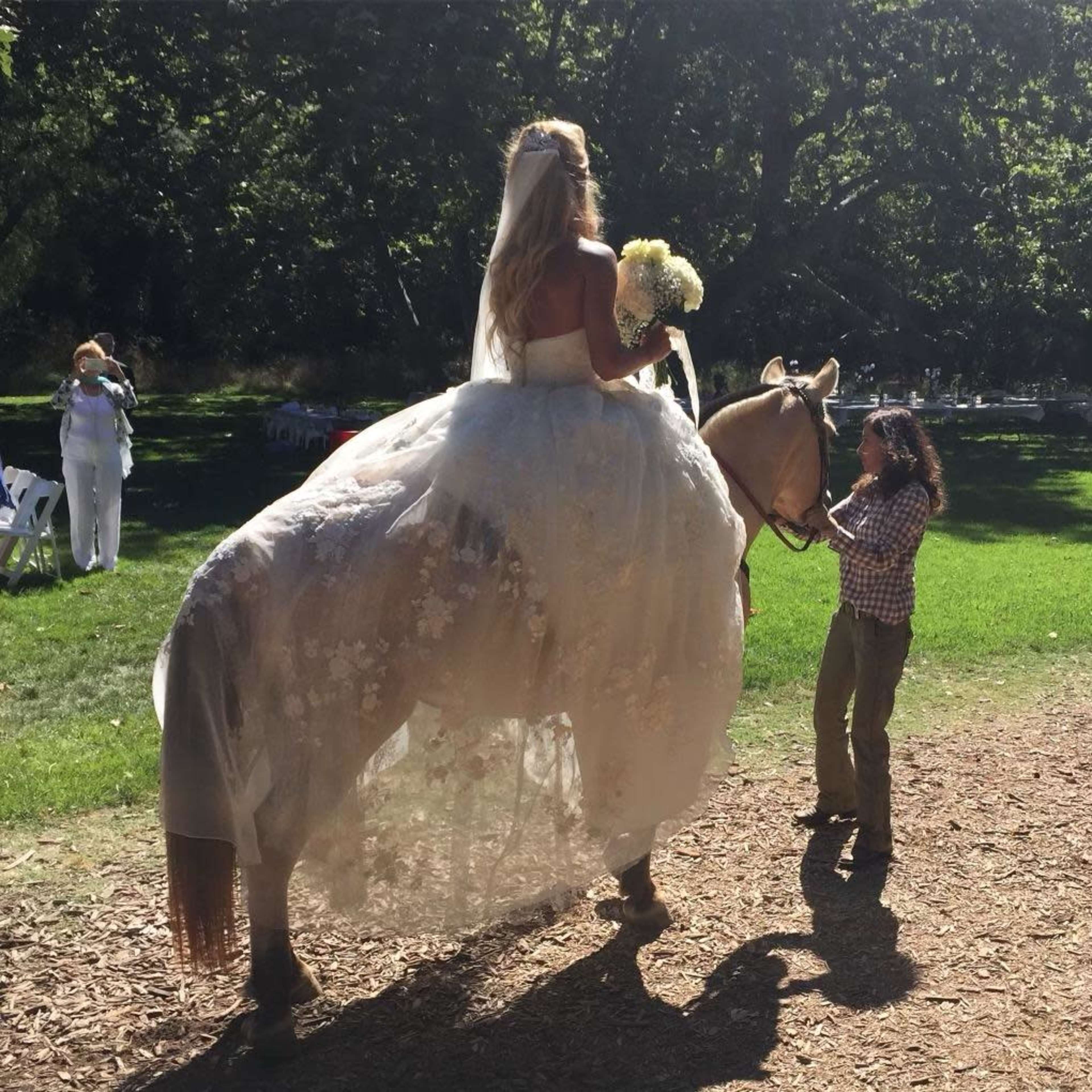 A bride in a white wedding dress stands atop a horse while holding a bouquet, with a person guiding the horse beside her.